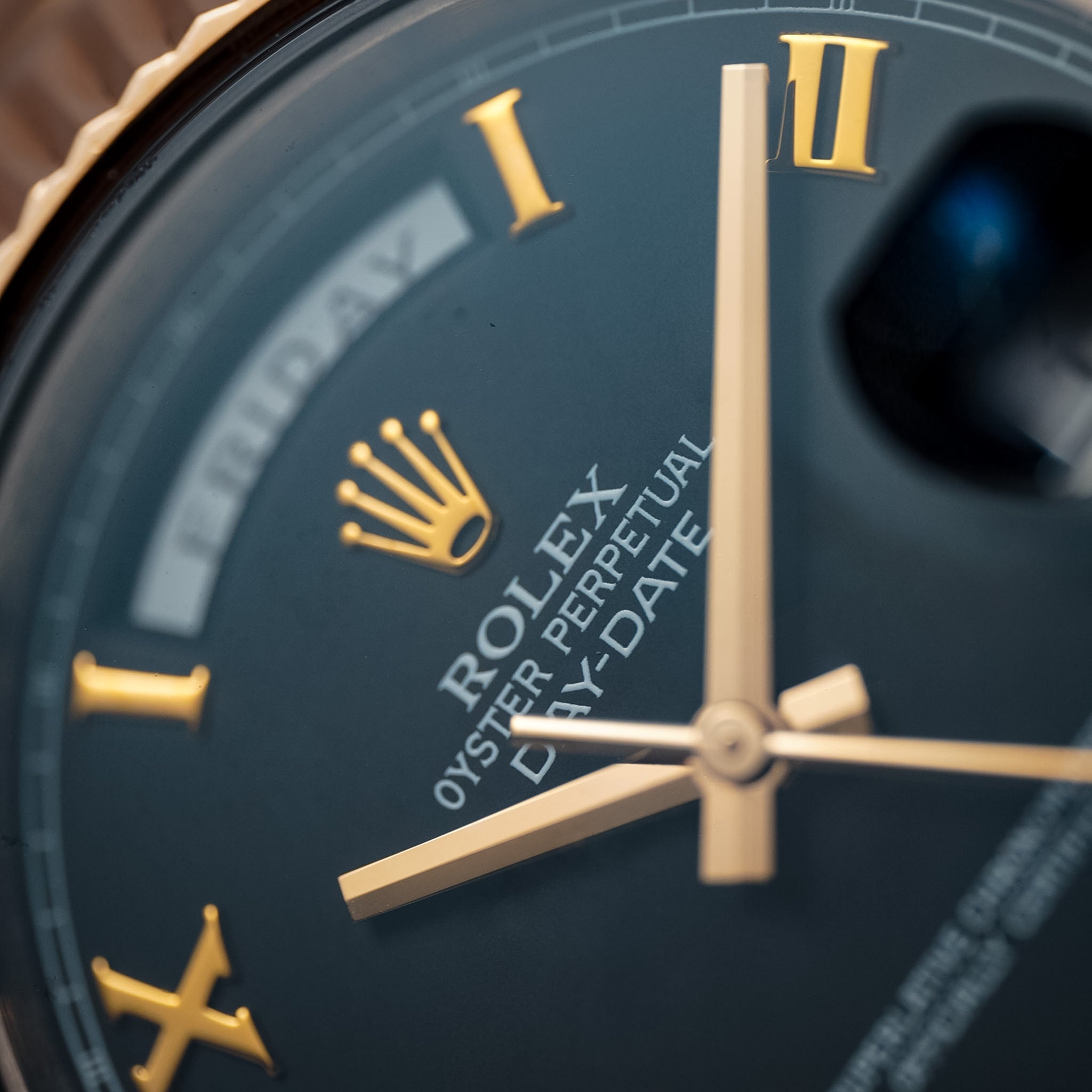 A close-up of a black dial watch with rose gold Roman numeral hour markers and a day indicator and Rolex logo at the top of the dial.