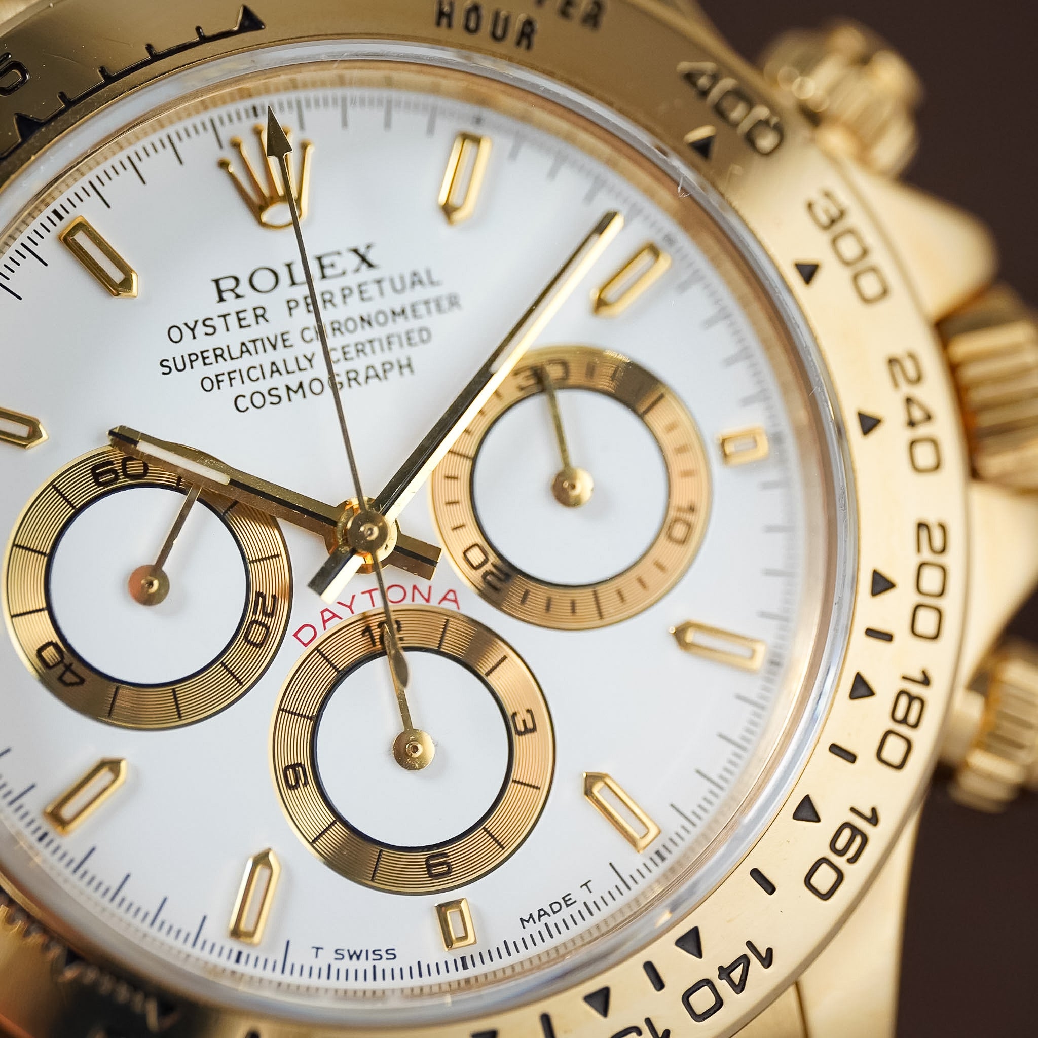 Close-up of a Rolex Daytona watch dial and bezel, showing a gold-tone case, white dial, gold hour markers and subdials, and a black tachymeter bezel.