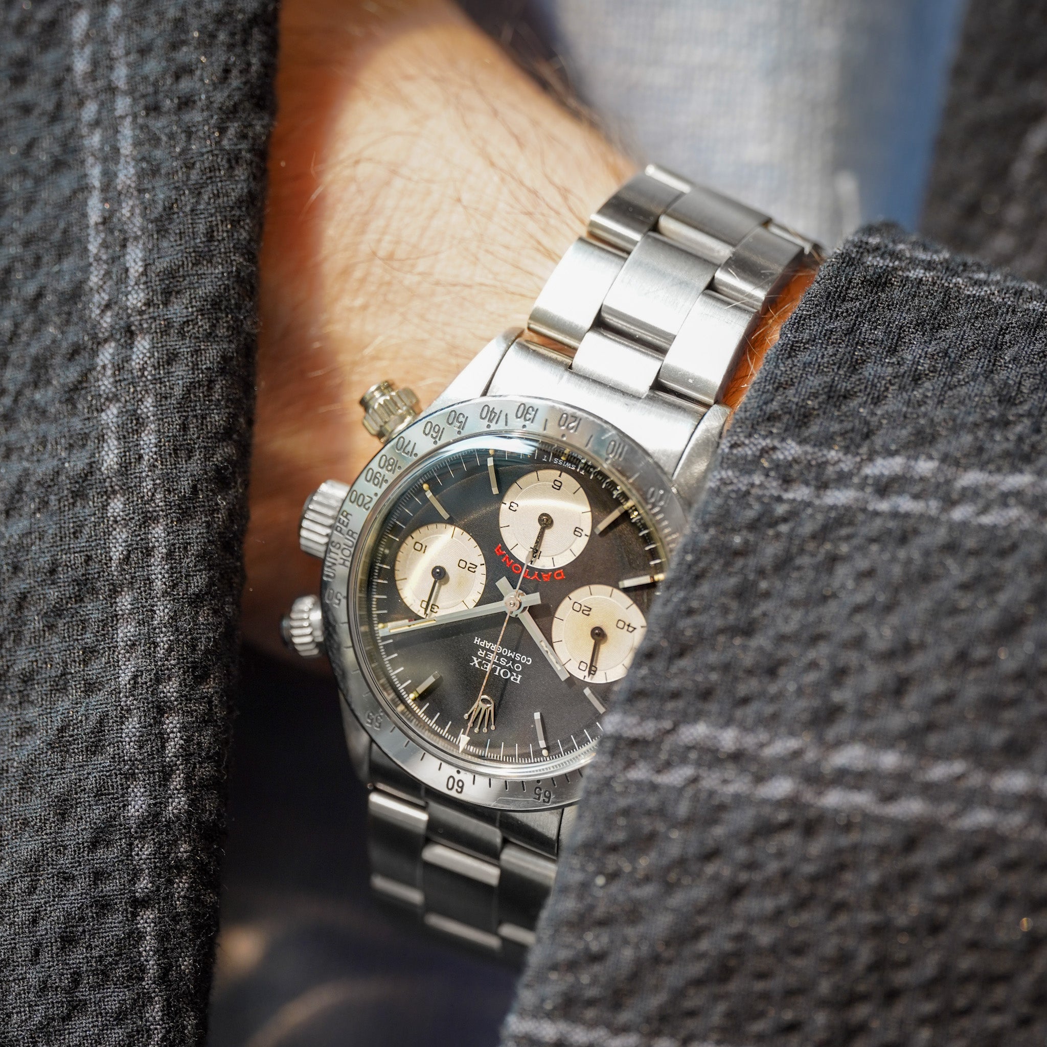 A close-up of a wrist wearing a silver stainless steel chronograph watch with a dark gray dial and beige subdials, framed by a gray textured jacket.