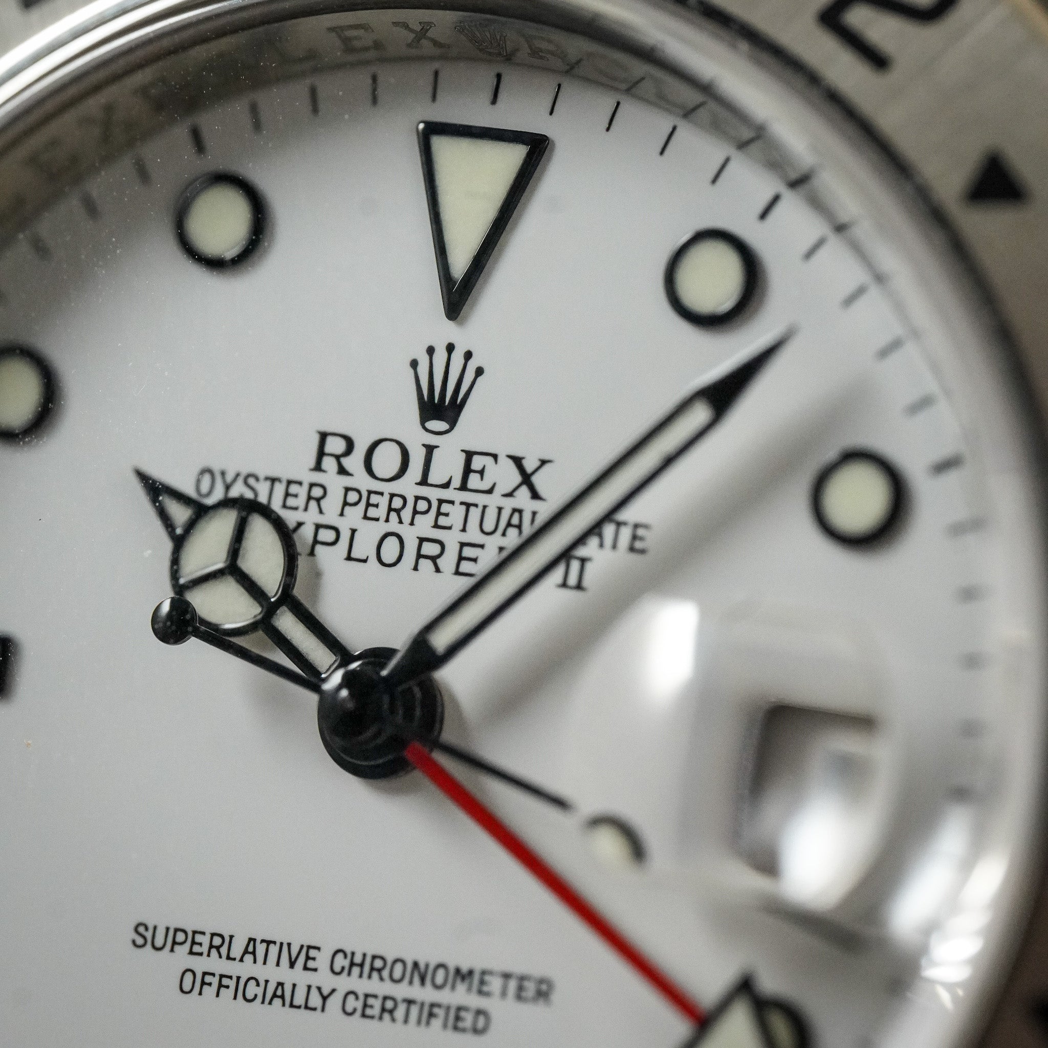 Close-up of a Rolex Oyster Perpetual Explorer II watch dial, showing the white face, black hands, luminous hour markers, crown logo, and a red seconds hand.
