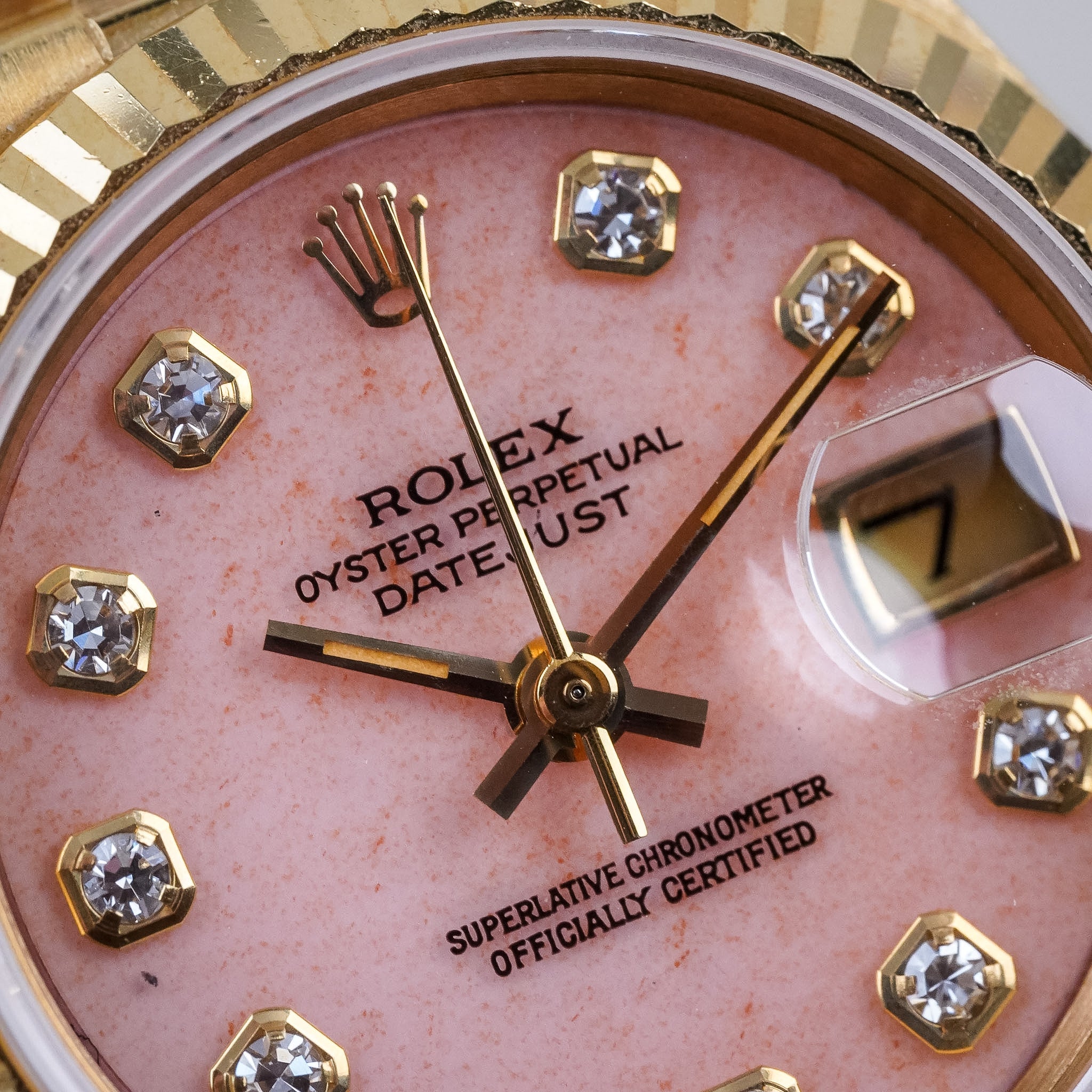 Closeup of a yellow gold Rolex Lady Datejust with a fluted bezel, pink coral dial, diamond hour markers, gold hands, and a date window at 3 o'clock showing "7".