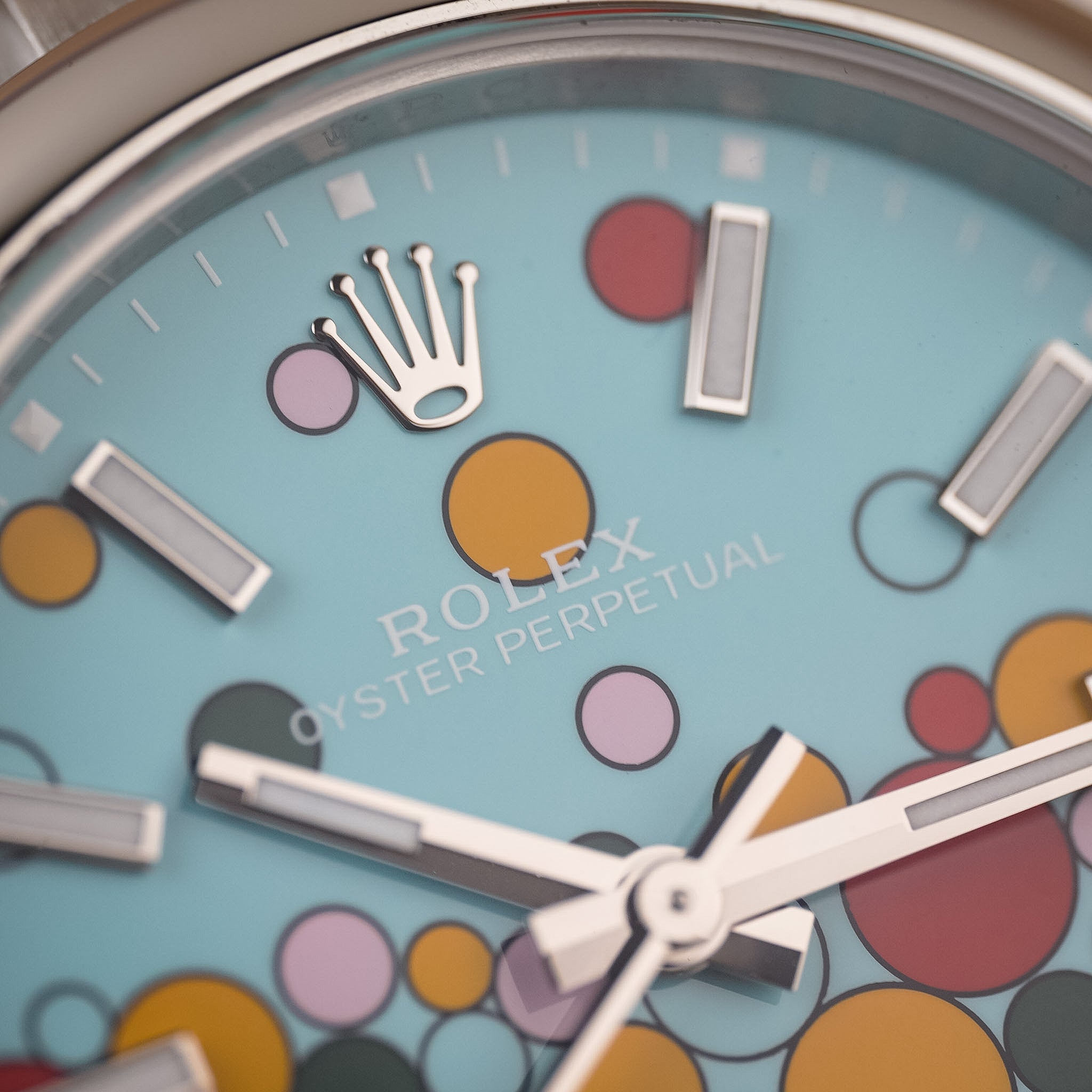 Closeup of a stainless steel Rolex watch with a colorful blue dial featuring accents of red, yellow, pink, and green. One can read the text Rolex Oyster Perpetual.