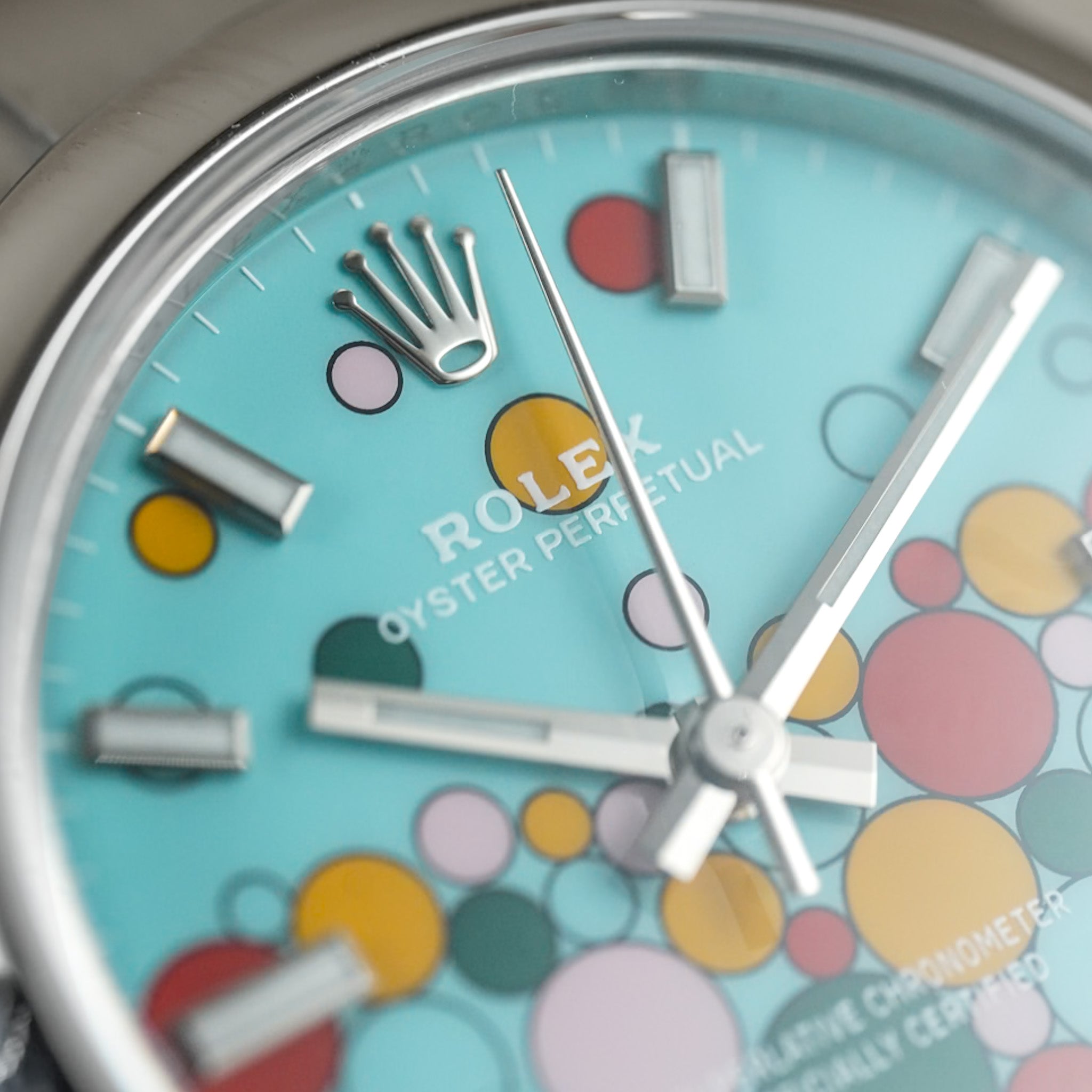 Close-up of a Rolex Oyster Perpetual watch dial with a light blue background and colorful bubble pattern in yellow, pink, green, and orange, with silver-tone hands and markers.