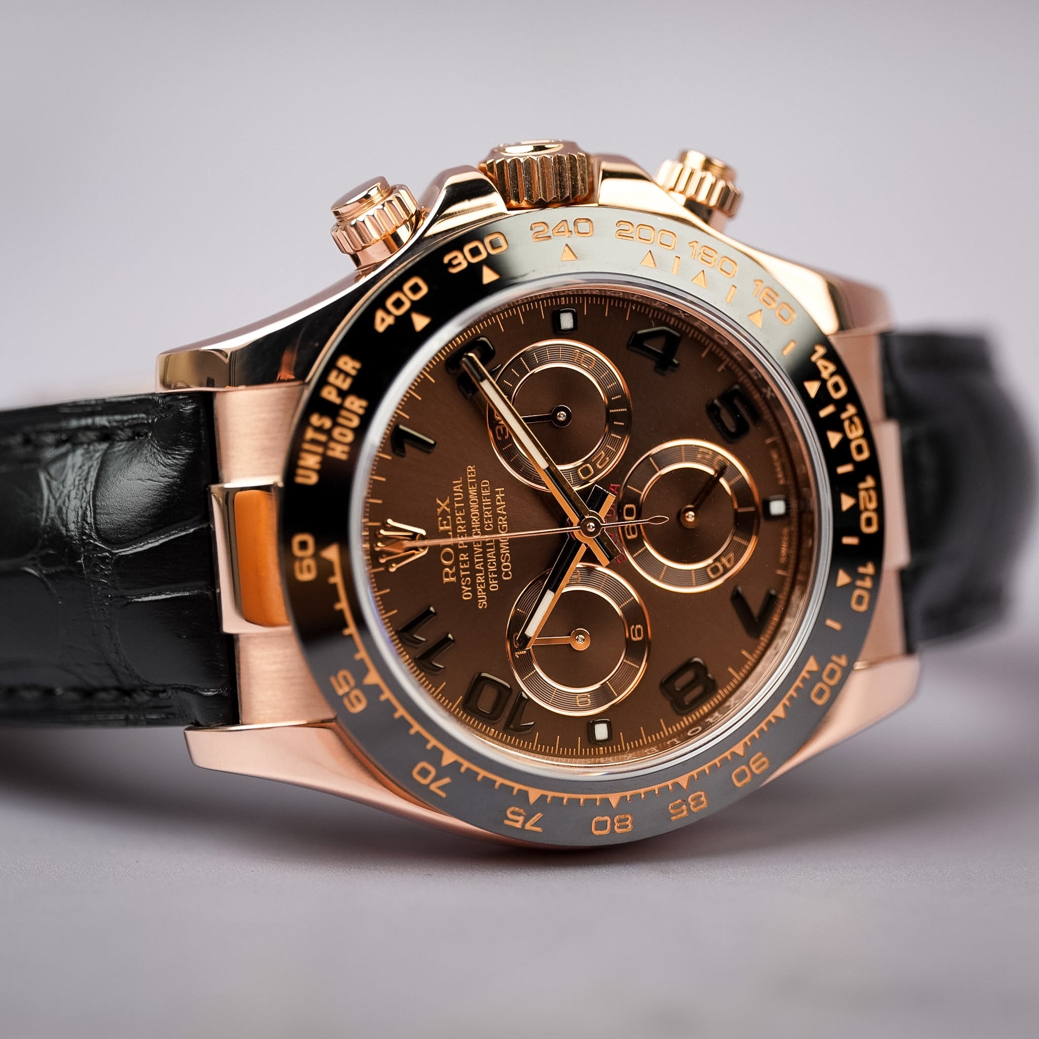 A rose gold Rolex Daytona with a chocolate brown dial, black ceramic bezel, and a black leather strap. Shot resting on its side on a neutral grey background.