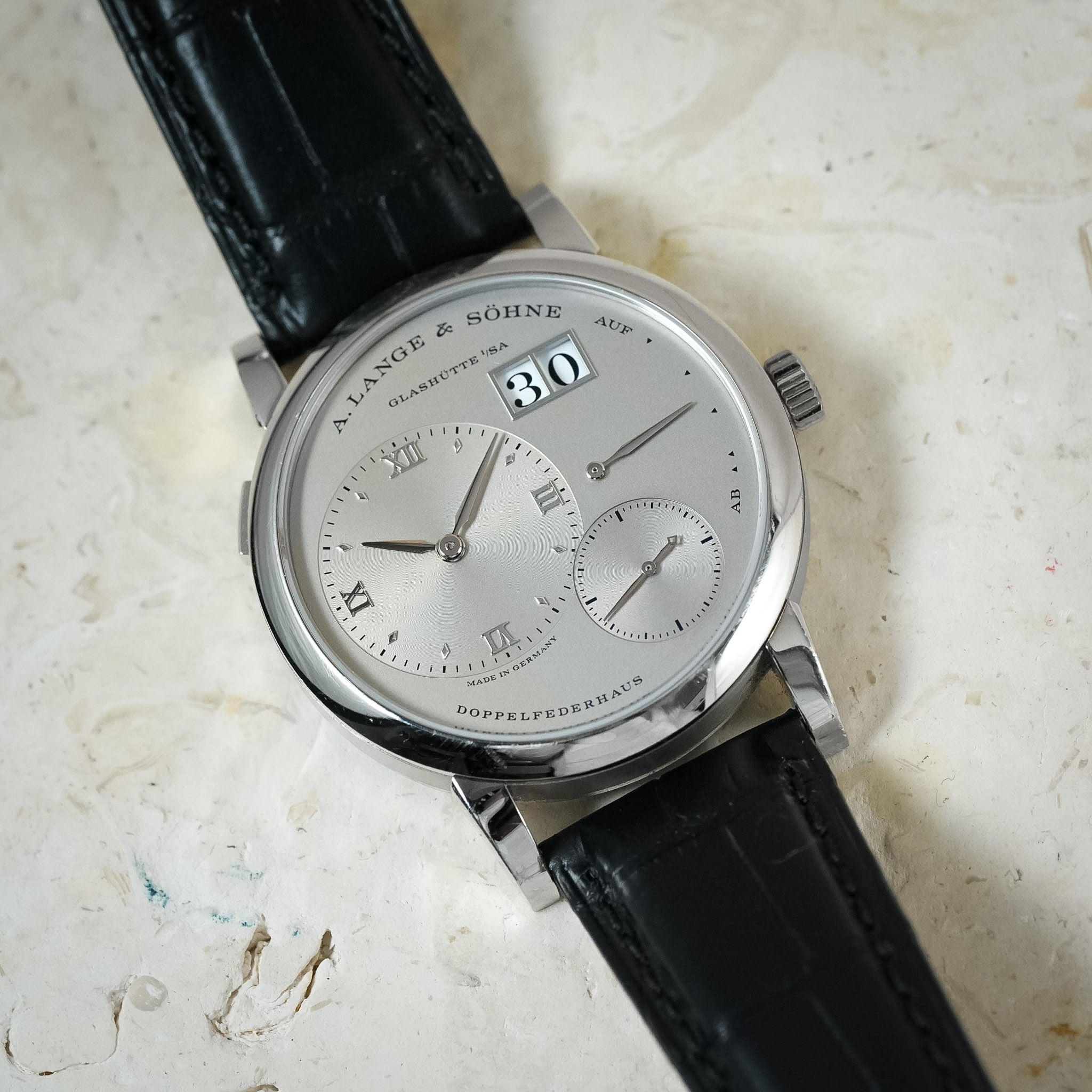 A close-up of a Lange & Söhne Quartzett-Schacht watch with a silver dial, Roman numeral hour markers, a date window showing 30, and a black leather strap on a textured off-white surface.