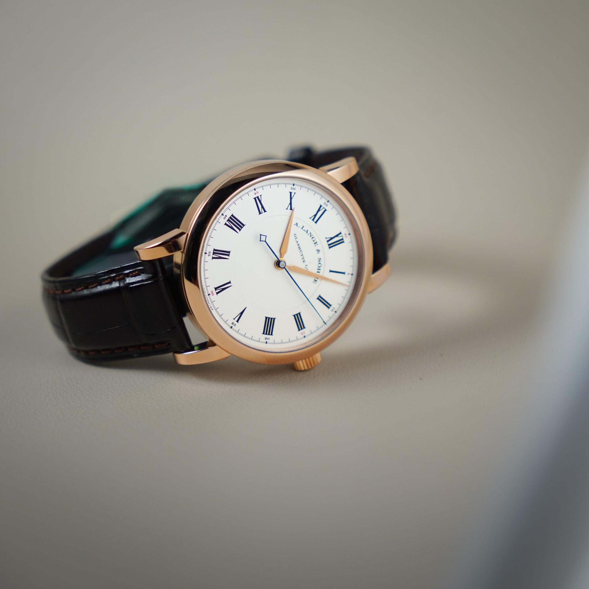 A luxury wristwatch with a rose gold case, white dial, blue markers and hands, and a black leather strap, resting on a neutral gray surface.