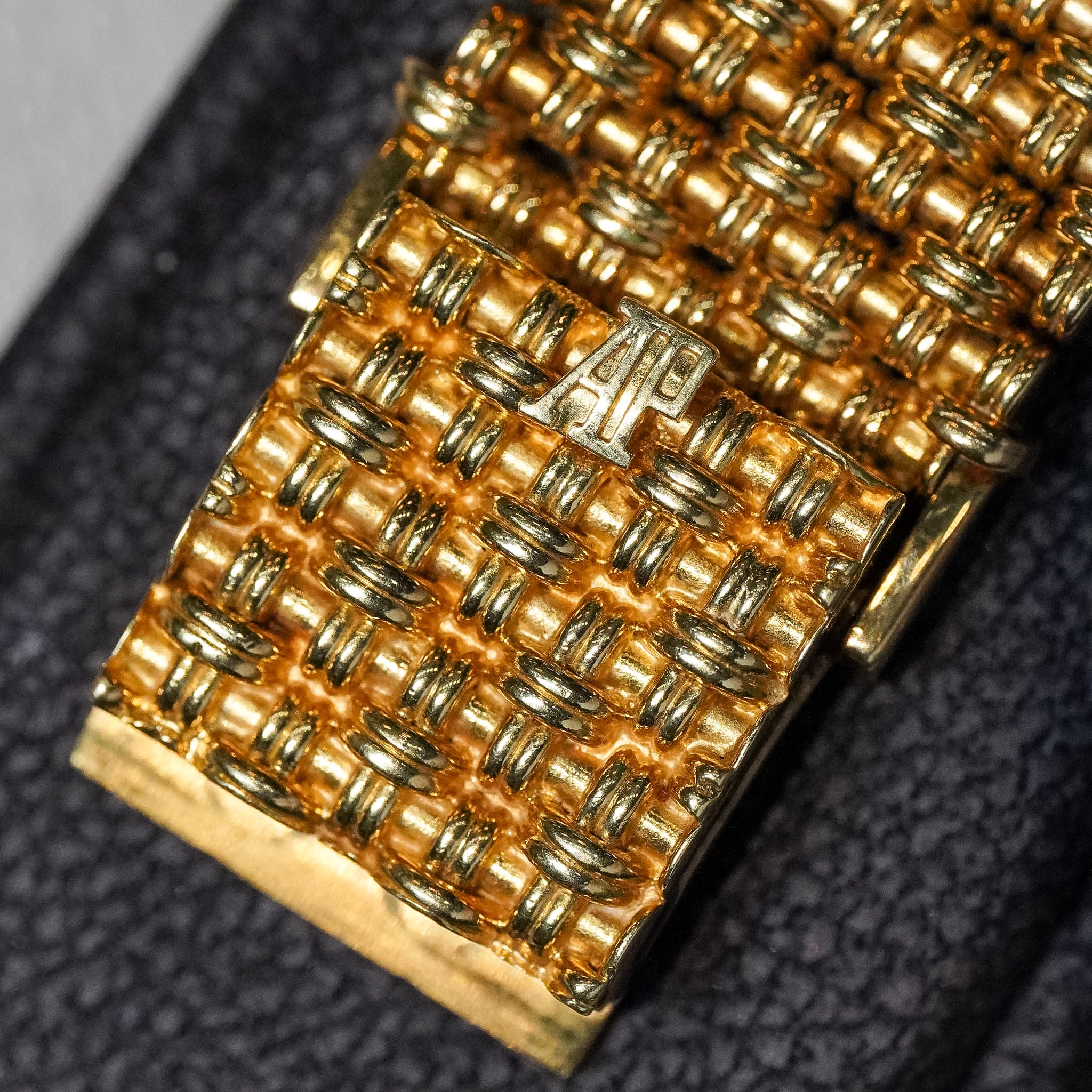 Close-up of a gold-toned woven bracelet or watch band with a small silver-toned AP logo plaque, resting on a dark textured surface.