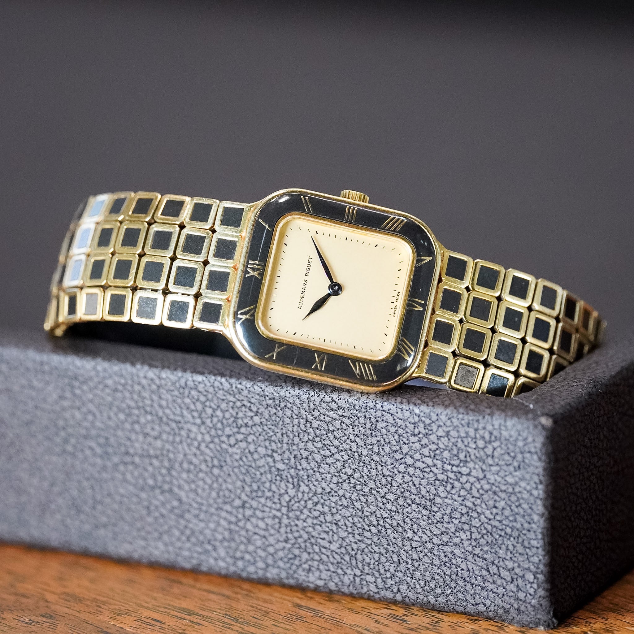 A vintage-style rectangular wristwatch with a gold-tone metal link bracelet and a cream-colored dial, resting on a textured gray display block.