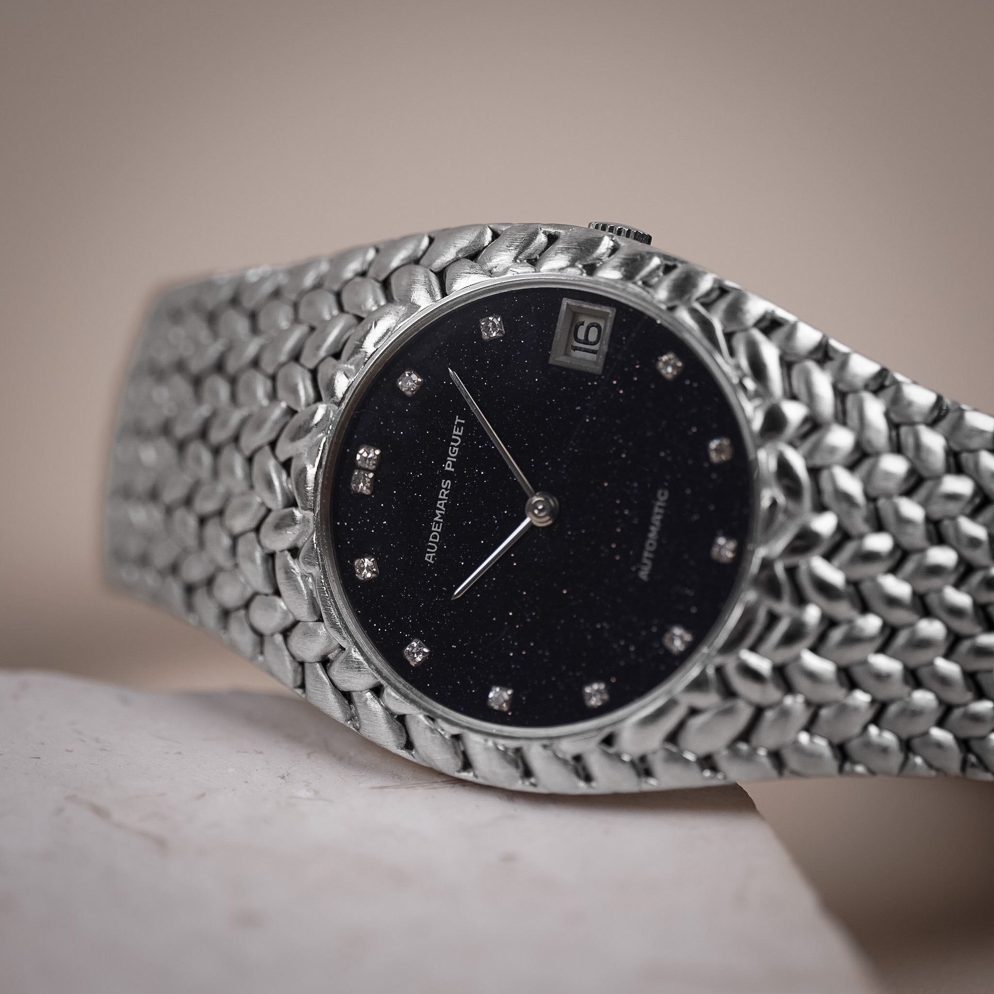 An Audemars Piguet wristwatch with a woven white gold case, diamond hour markers, date window, and an aventurine dial sits diagonally on a light grey stone slab.