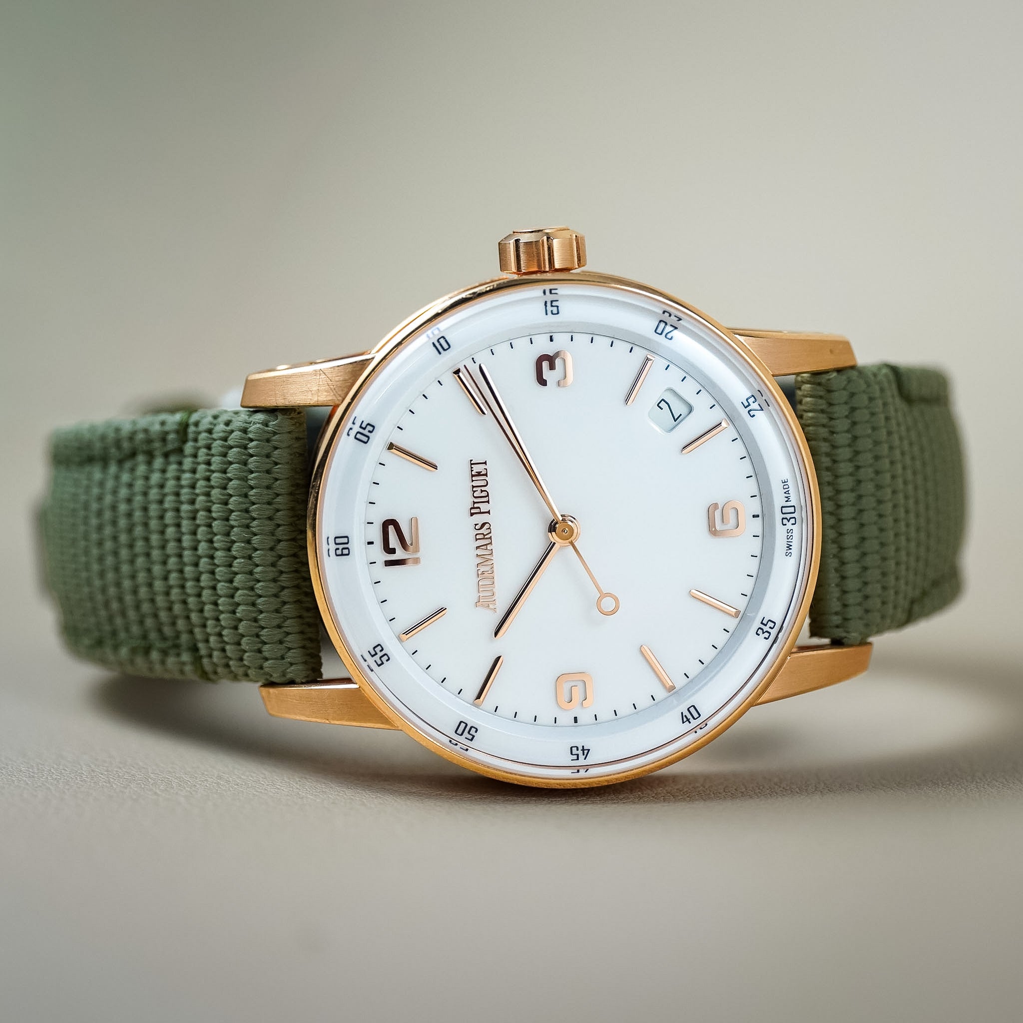 A wristwatch with a white dial, rose gold case, and an olive green fabric strap, shown on a neutral beige background.