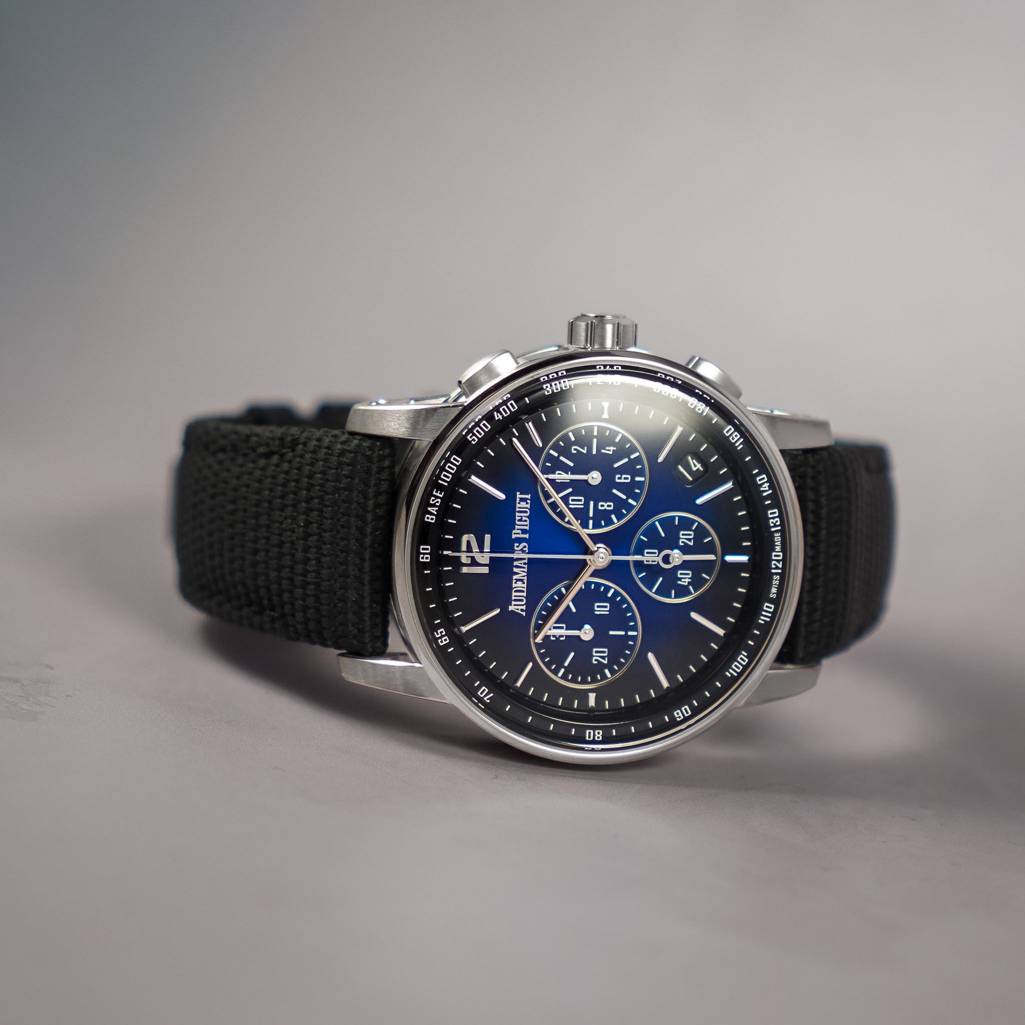 A wristwatch with a silver-tone round case, a dark blue chronograph dial with white markings and subdials, and a black woven fabric strap, photographed on a neutral gray background.