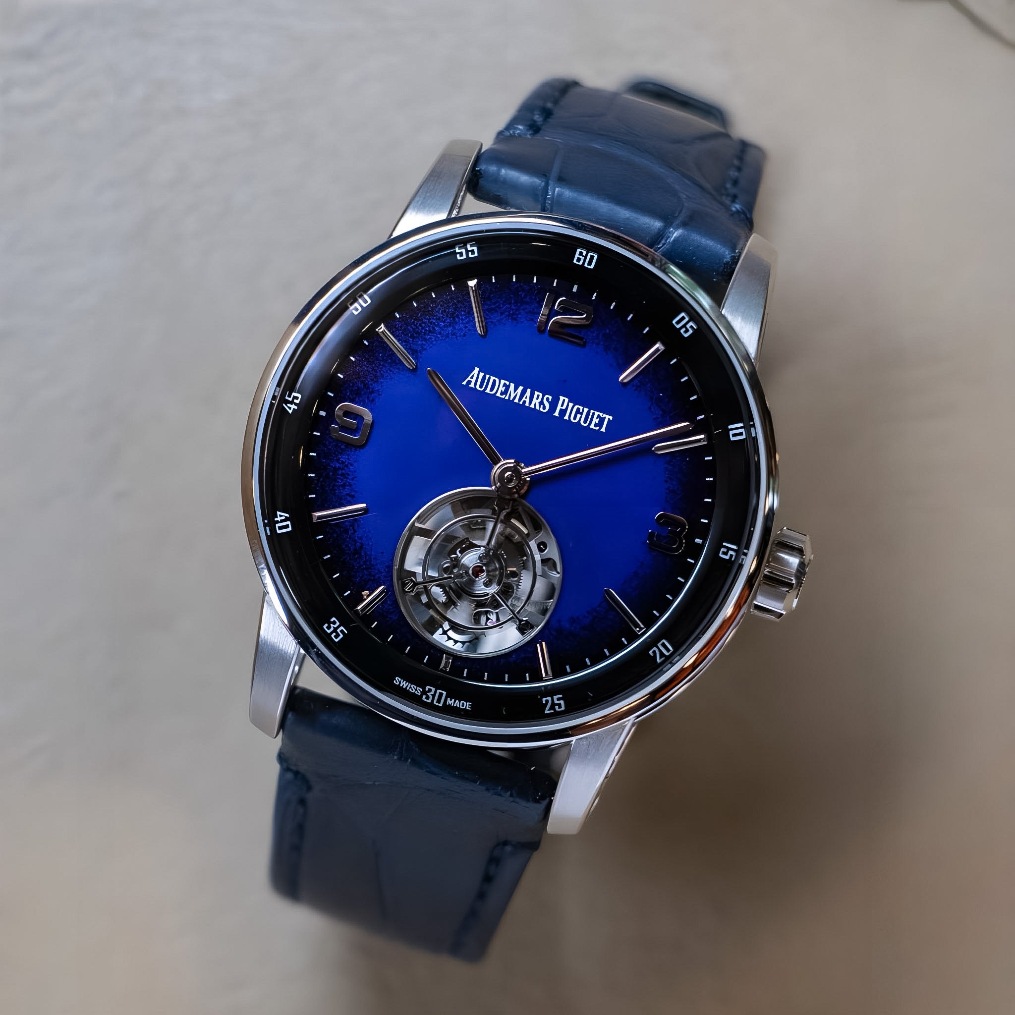 Audemars Piguet Royal Oak watch featuring a vibrant blue sunburst dial, a visible tourbillon under the dial, and a dark blue leather strap, shown on a neutral beige background.