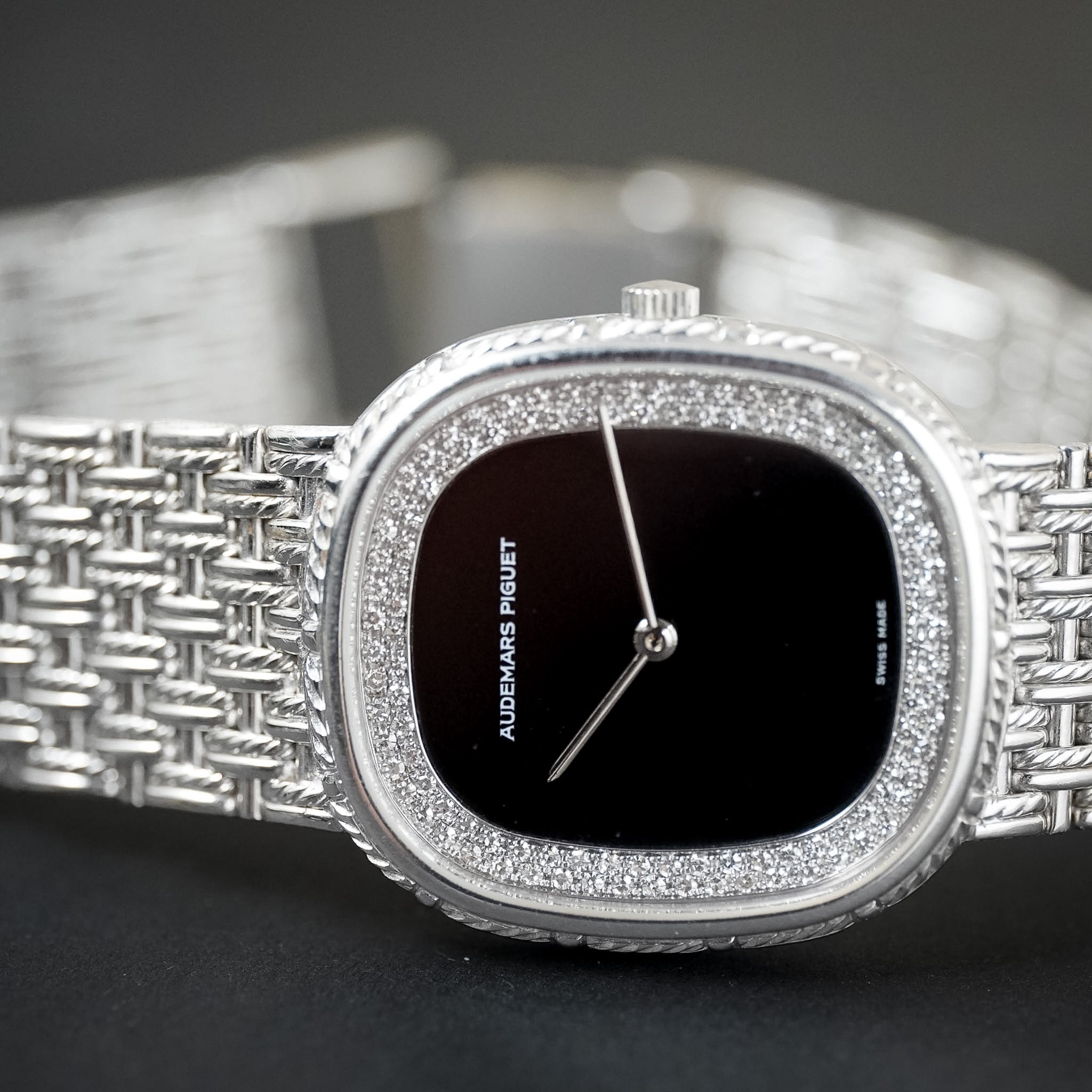 A close-up of a silver-tone Audemars Piguet watch with a textured woven bracelet, a diamond-encrusted bezel, and a black dial.