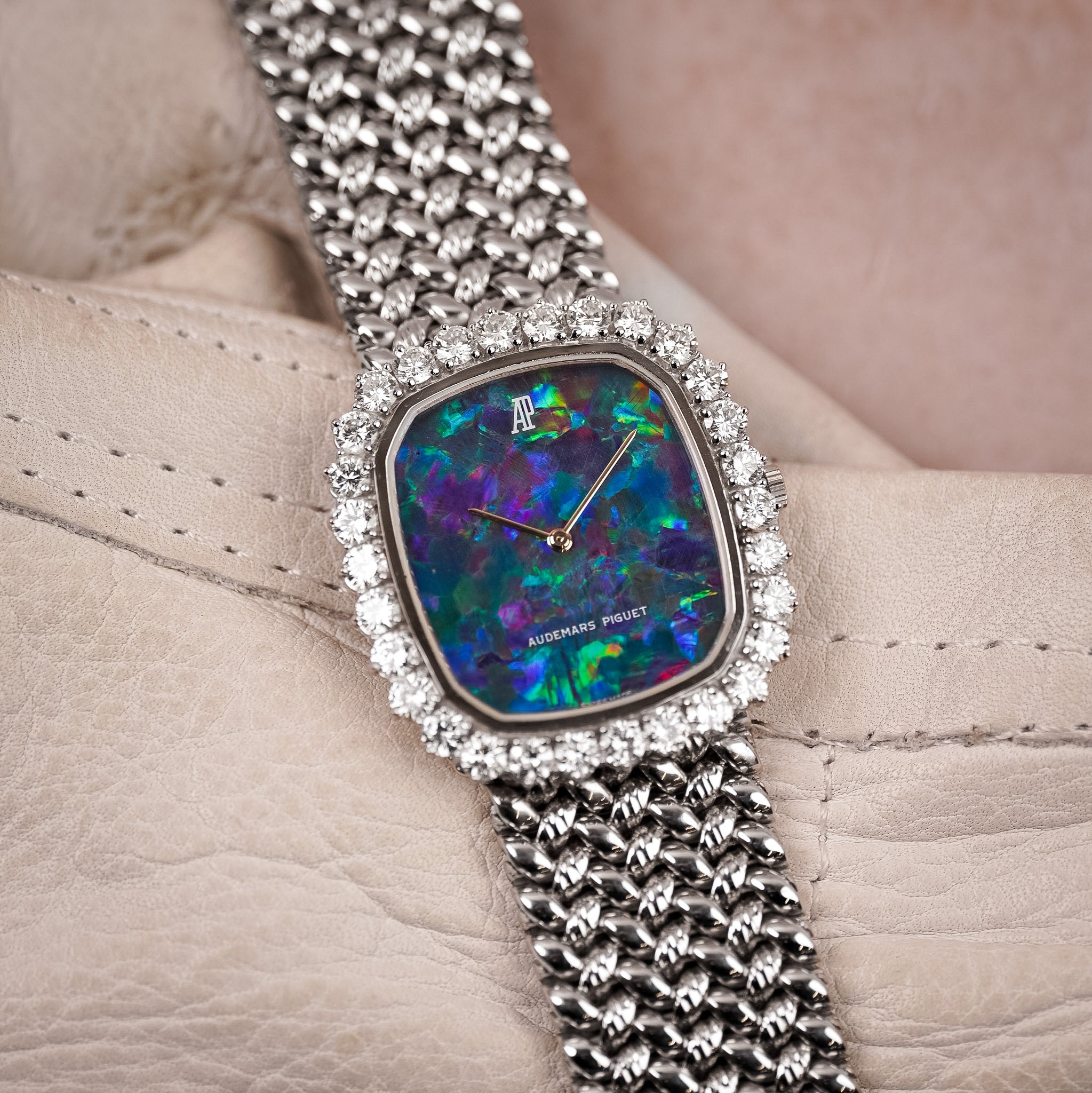 A luxury women's watch by Audemars Piguet, featuring a square bezel set with clear crystals and a vibrant iridescent mother-of-pearl dial. The watch has a silver-toned mesh bracelet and is displayed on a beige leather surface.