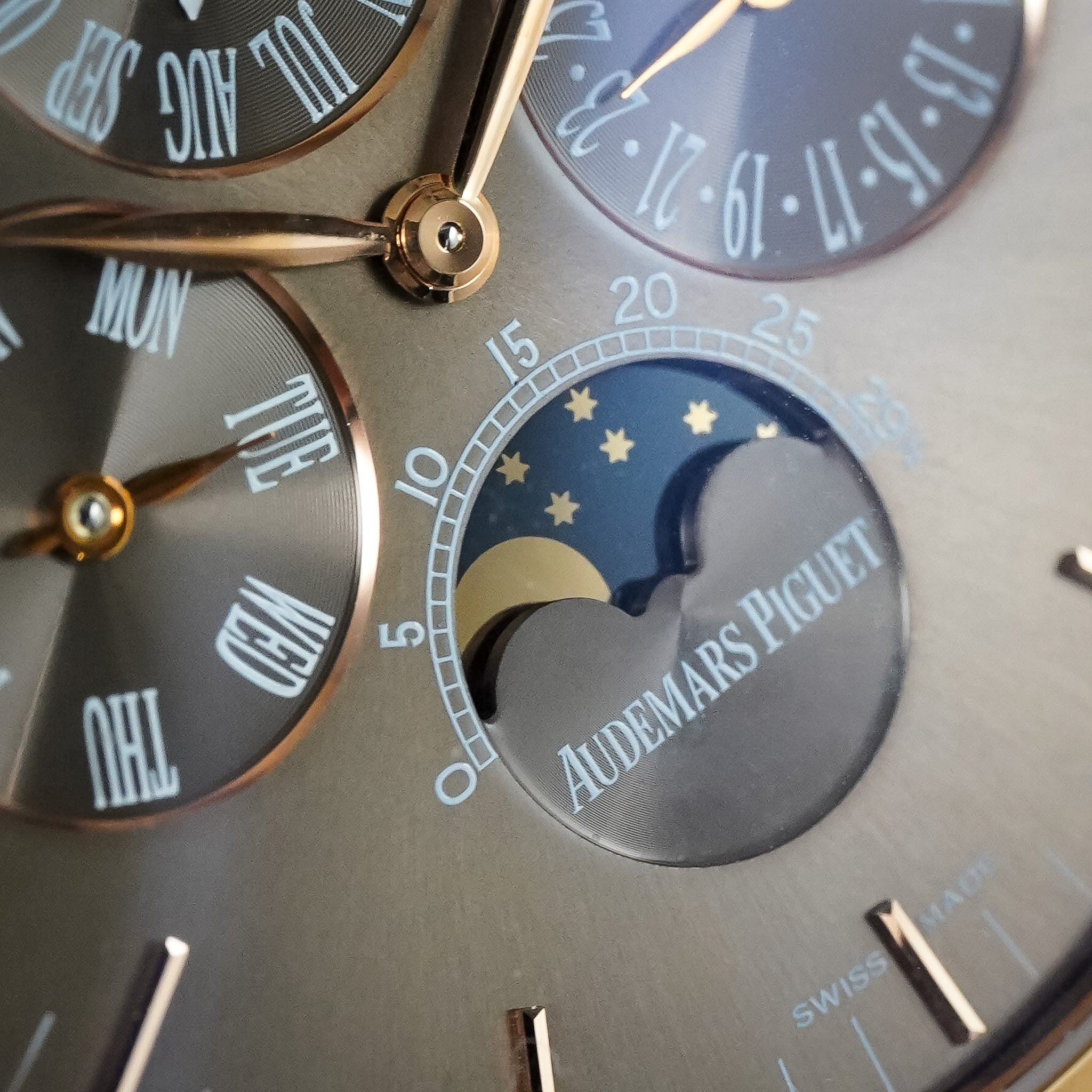 Close-up of an Audemars Piguet watch dial showing multiple subdials, including a moonphase window with stars and a crescent moon, in polished gold-tone metal on a taupe-gray case.