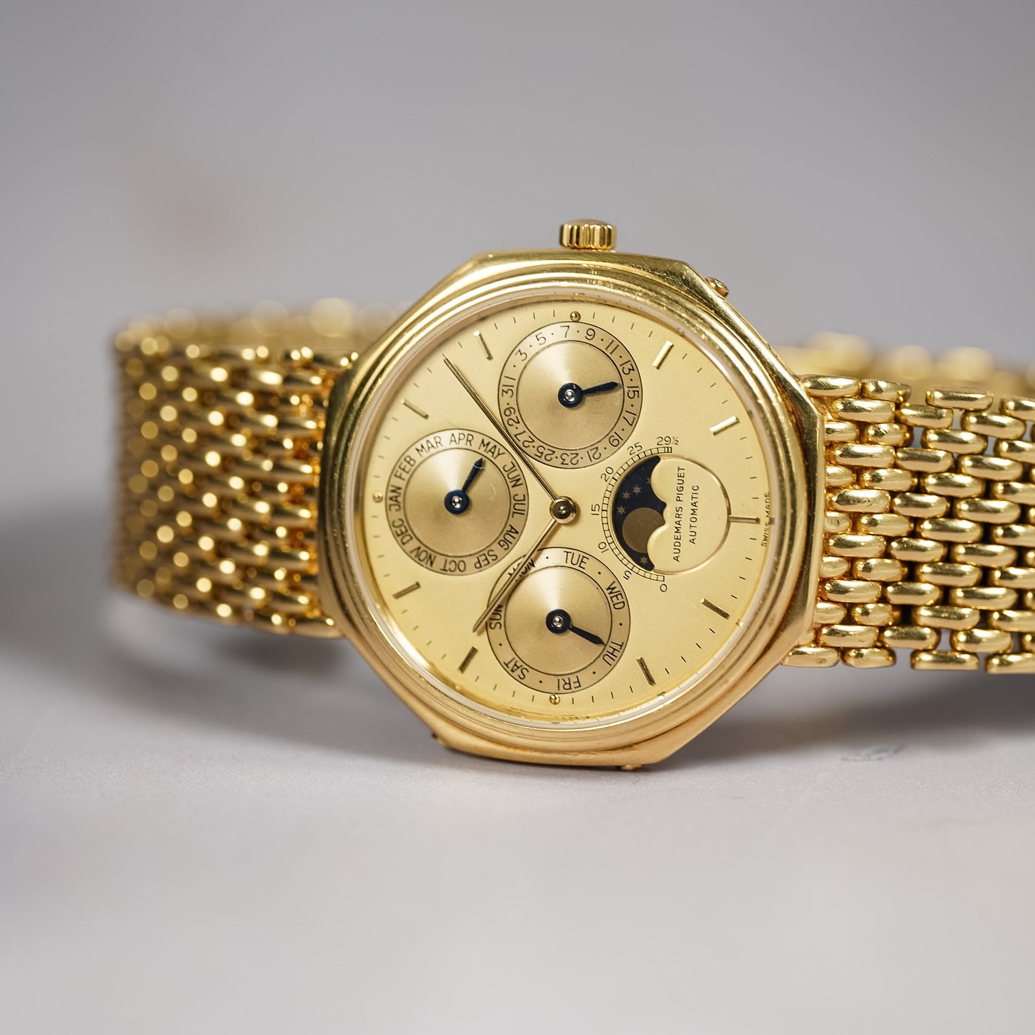 A gold-tone automatic watch with an octagonal bezel, cream-colored dial featuring three subdials and a moon phase window, paired with a matching gold mesh bracelet.