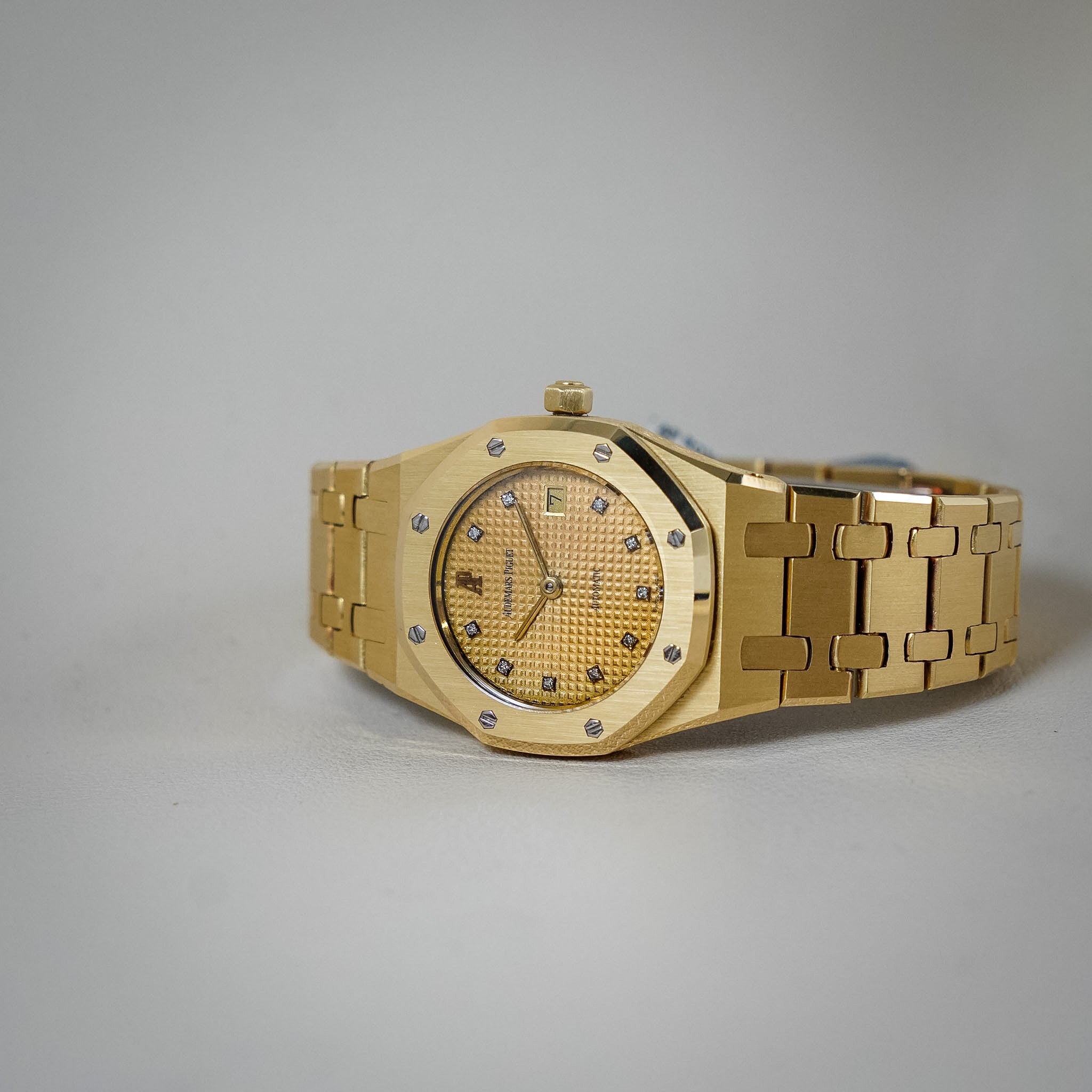 A gold wristwatch with an octagonal bezel, textured gold dial, and matching gold-tone link bracelet, photographed on a plain light gray background.