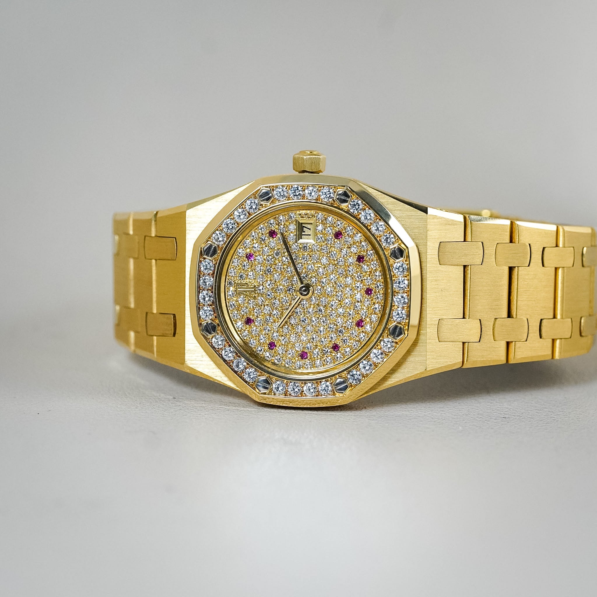 A luxury gold wristwatch with a diamond-encrusted bezel and a white diamond pavé dial accented with small red gemstones, shown on a neutral gray background.