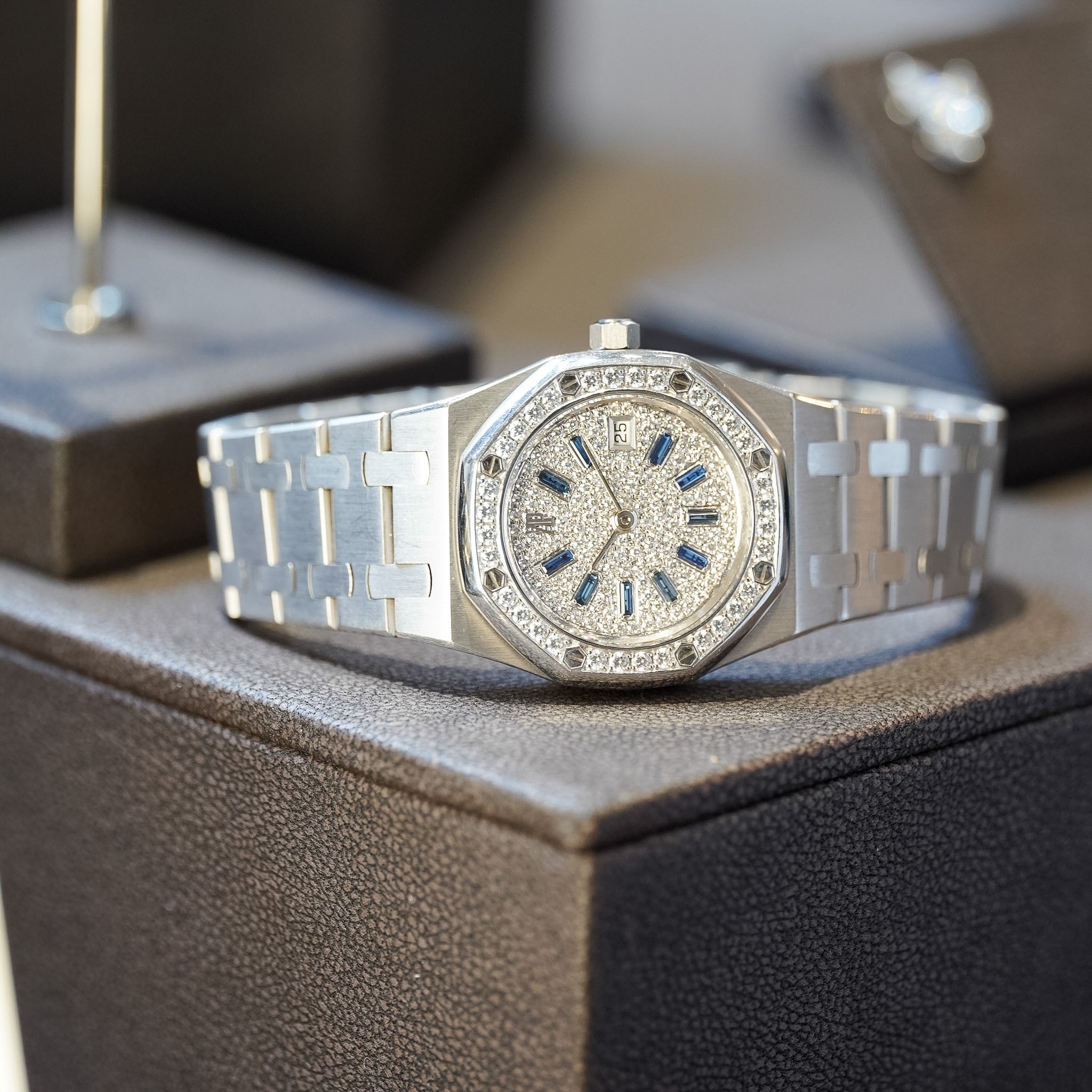 A silver-toned stainless steel wristwatch with a diamond-encrusted bezel, octagonal case, and matching link bracelet, resting on a textured dark brown watch box.