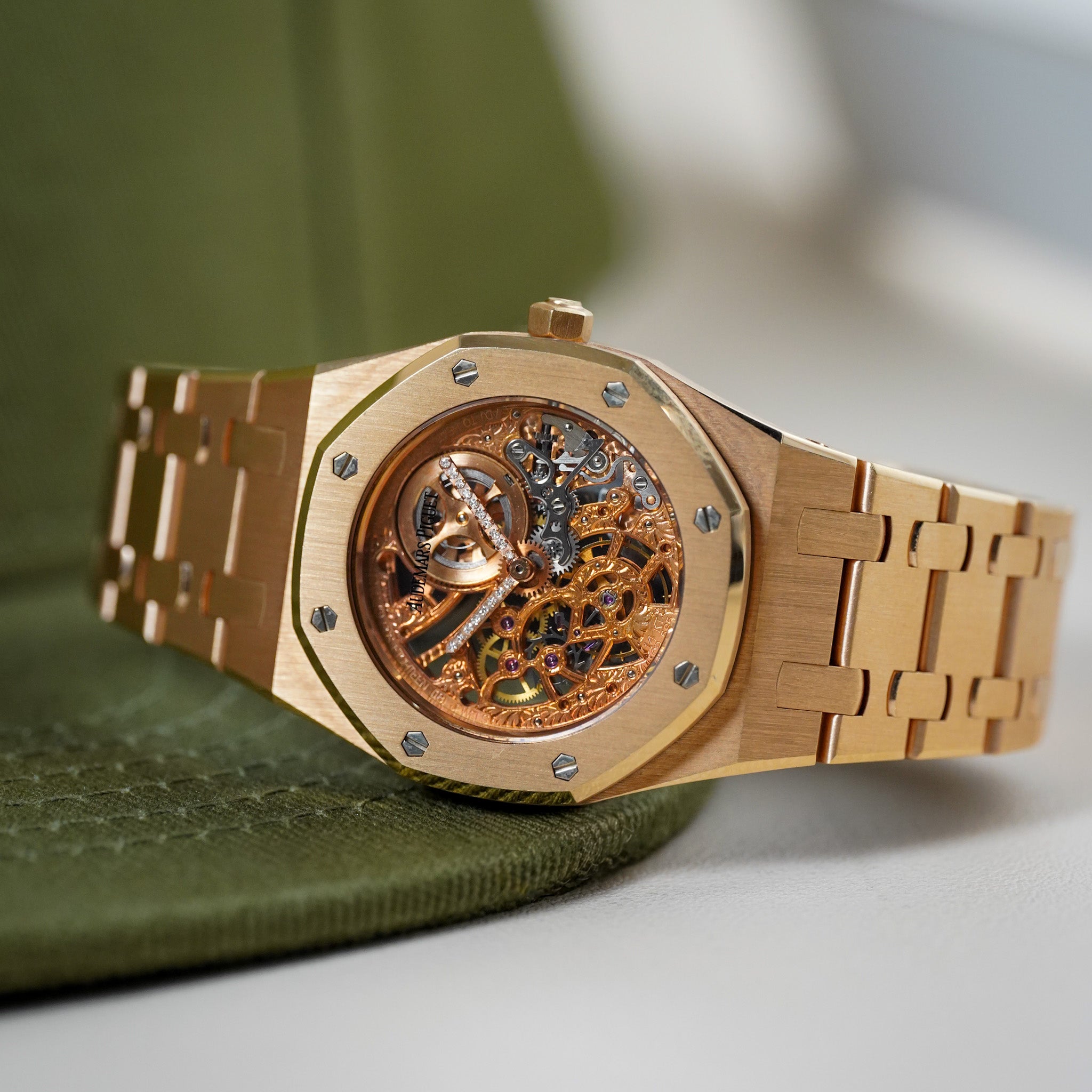 A gold-tone automatic watch with a skeleton dial showing intricate inner gears, resting on an olive green textured surface against a soft-focus light gray background.