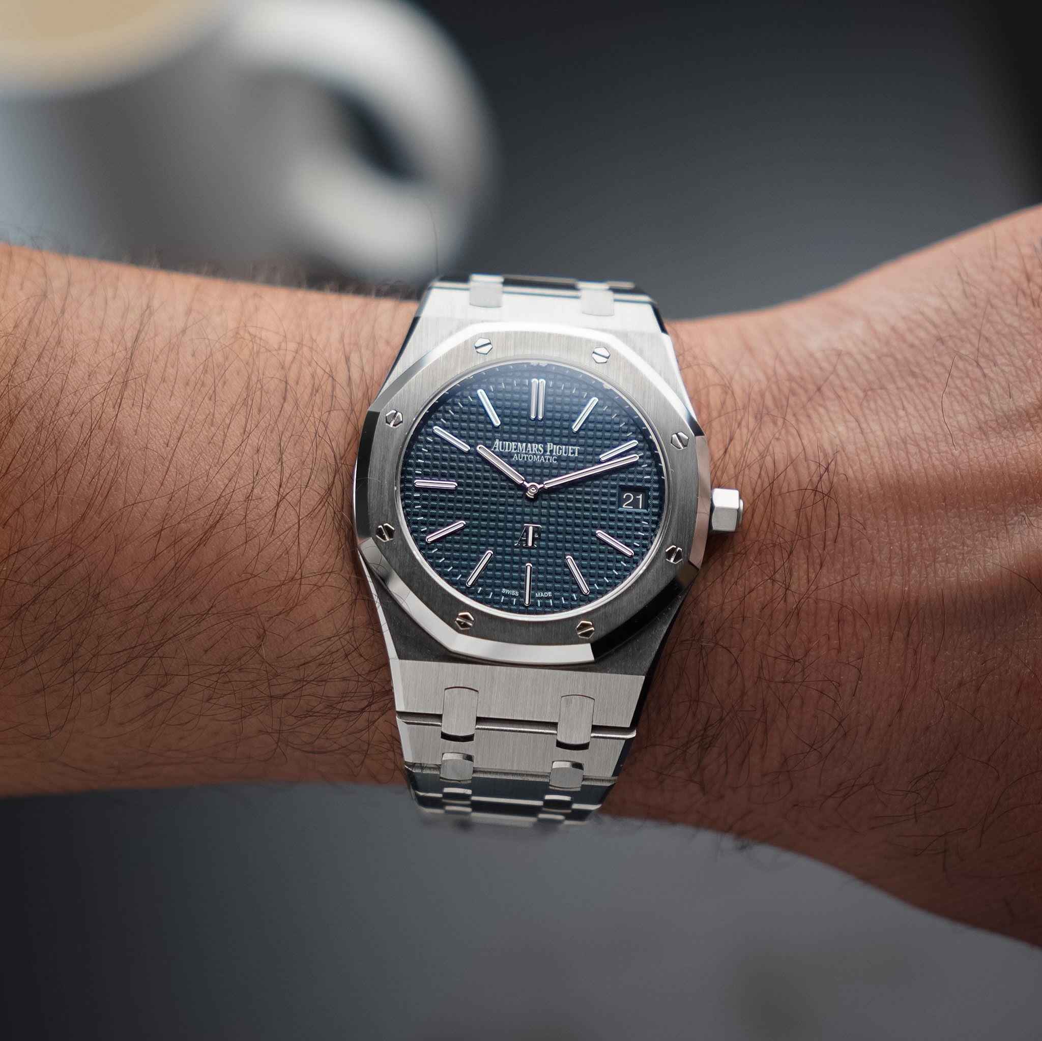 A steel Audemars Piguet Royal Oak with a steel bezel, textured blue dial with a date window, and steel bracelet, shot on a man's wrist with a blurry background.