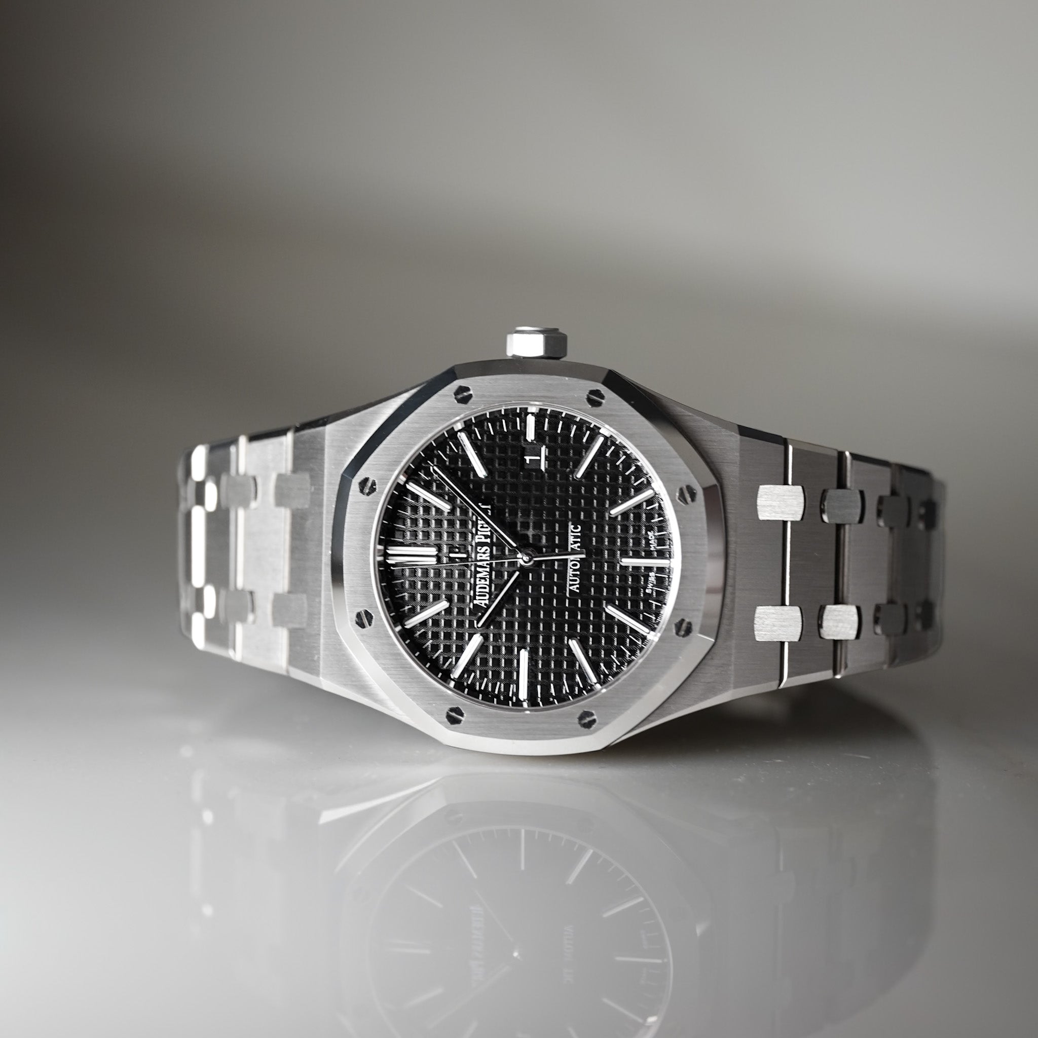 A luxury stainless steel wristwatch with an octagonal bezel, black textured dial, and matching steel link bracelet, displayed on a reflective gray surface.