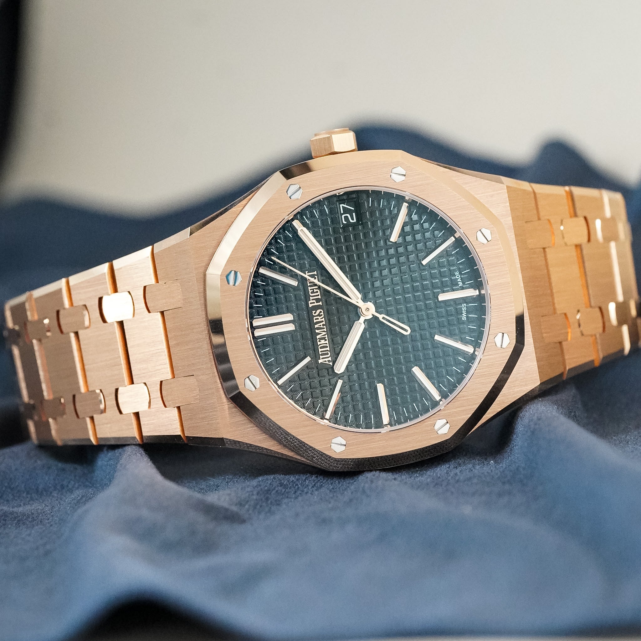 A rose gold Audemars Piguet wristwatch lies on its side. The watch has a textured black dial with rose gold hands and hour markers. It's sitting on black fabric. 