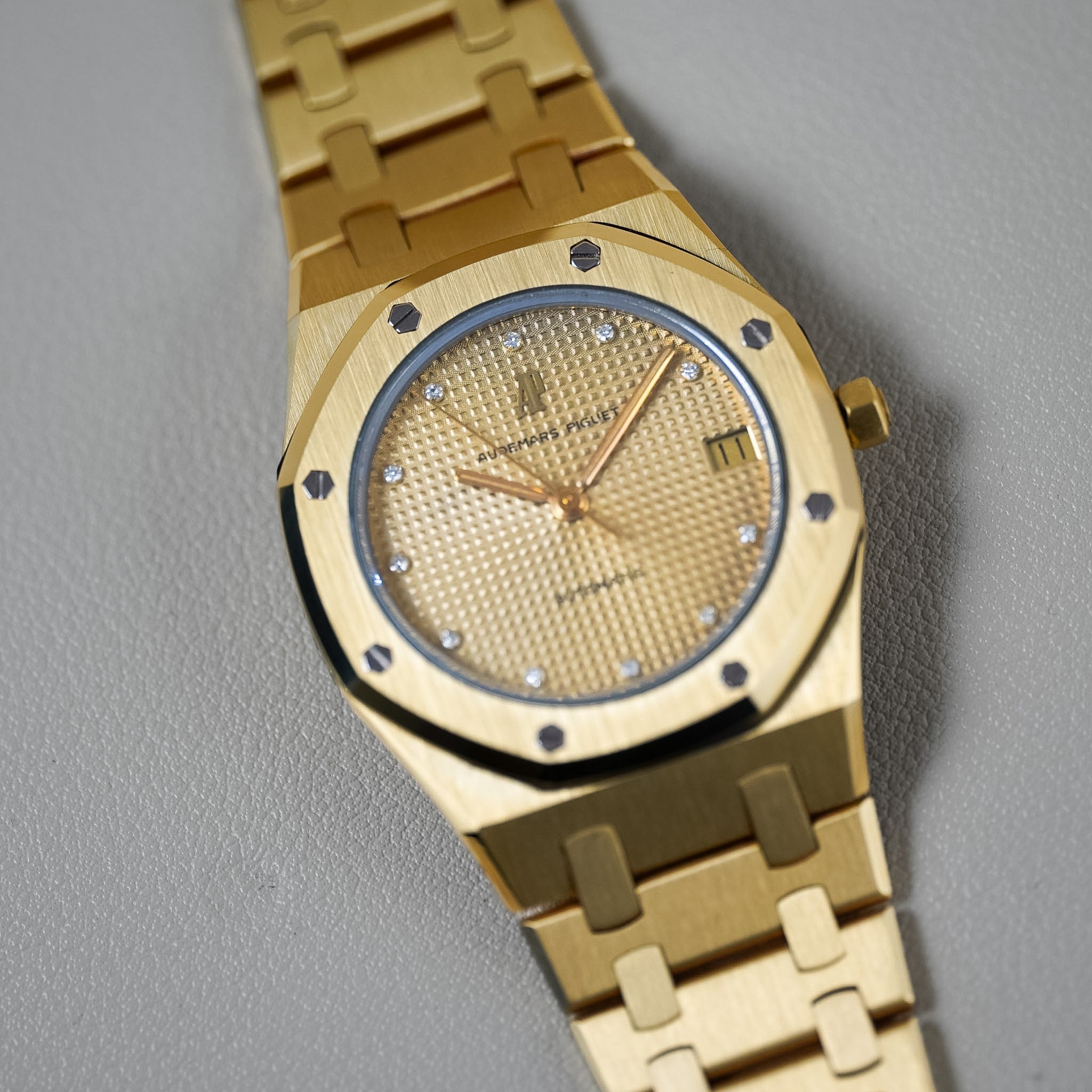 A gold watch with a textured dial, diamond hour markers, and a matching gold-tone link bracelet, photographed on a neutral gray background.
