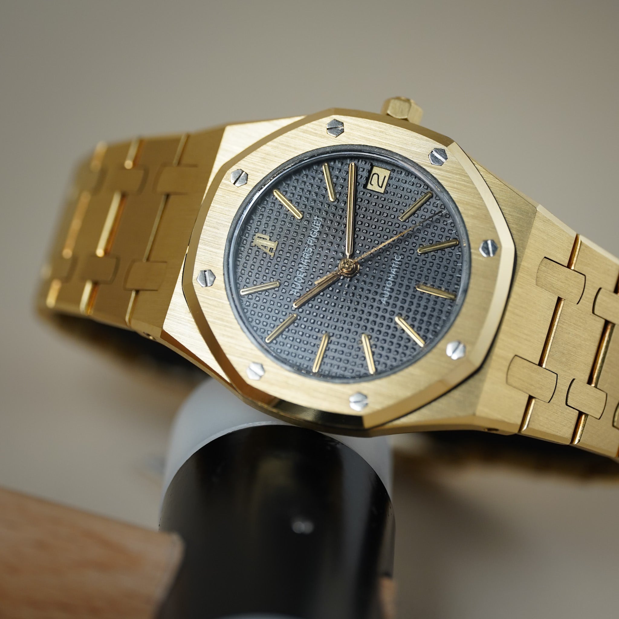 A gold watch with an octagonal bezel, gold-tone bracelet, and a textured dark gray dial, shown close-up resting on a black stand.