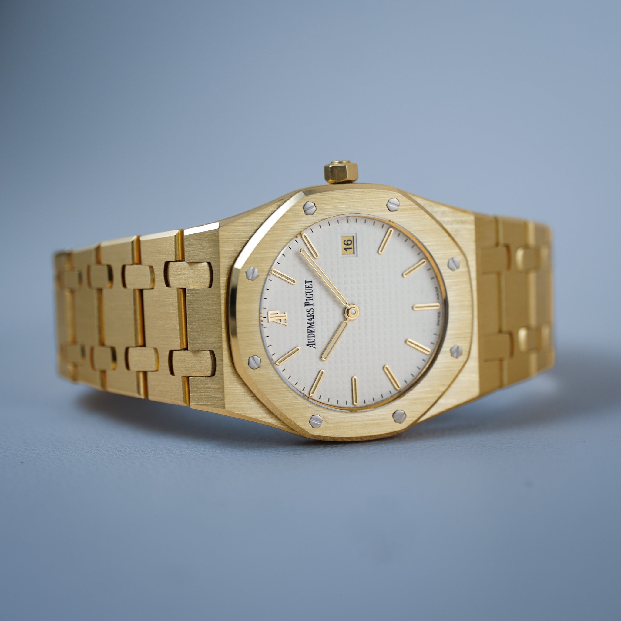 A gold-tone Audemars Piguet Royal Oak watch with an octagonal bezel, white dial, and matching gold-tone link bracelet, photographed on a clean light blue-gray background.