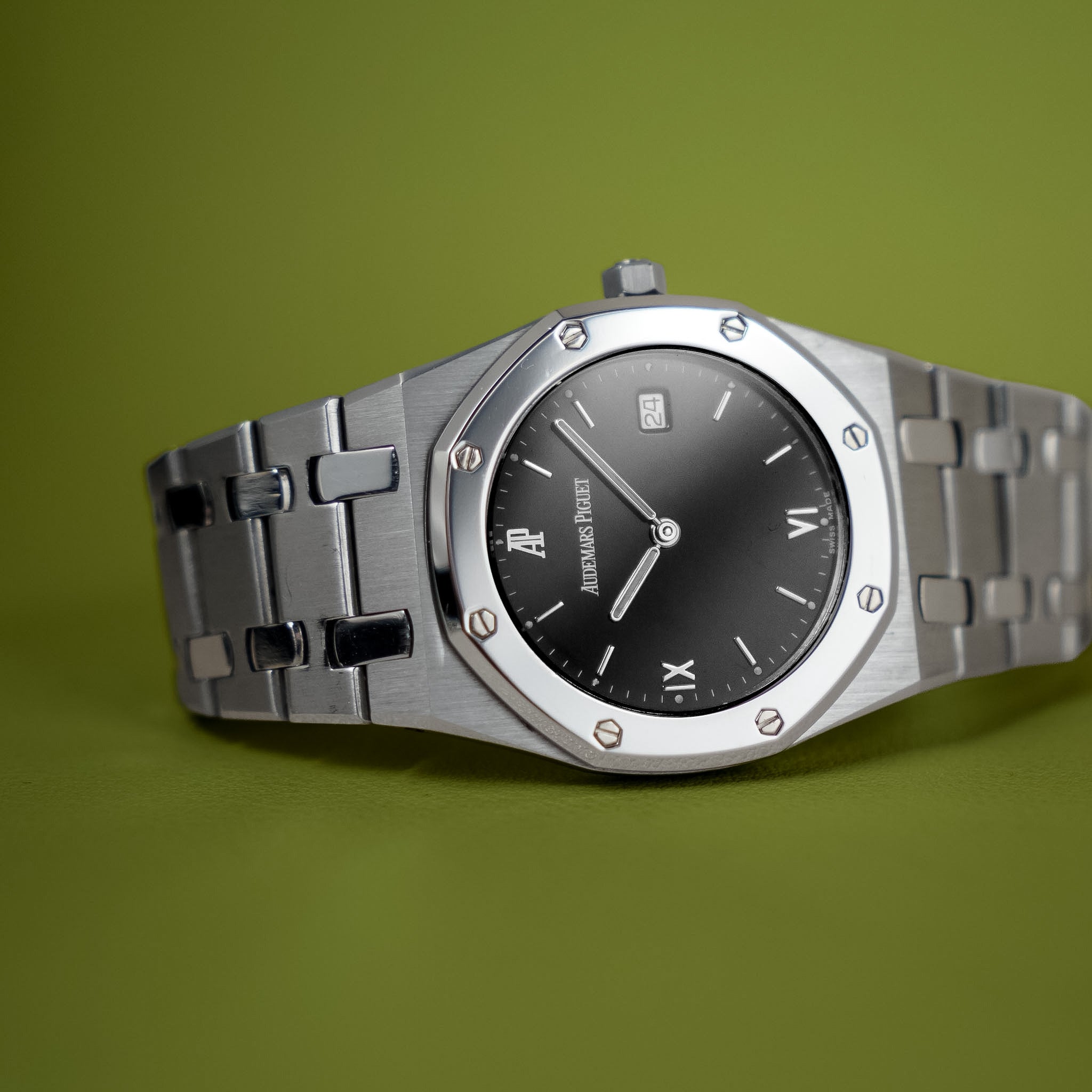 A stainless steel Audemars Piguet Royak Oakwristwatch with a black dial, Roman numeral hour markers, and a date window shot on a vibrant green background. 