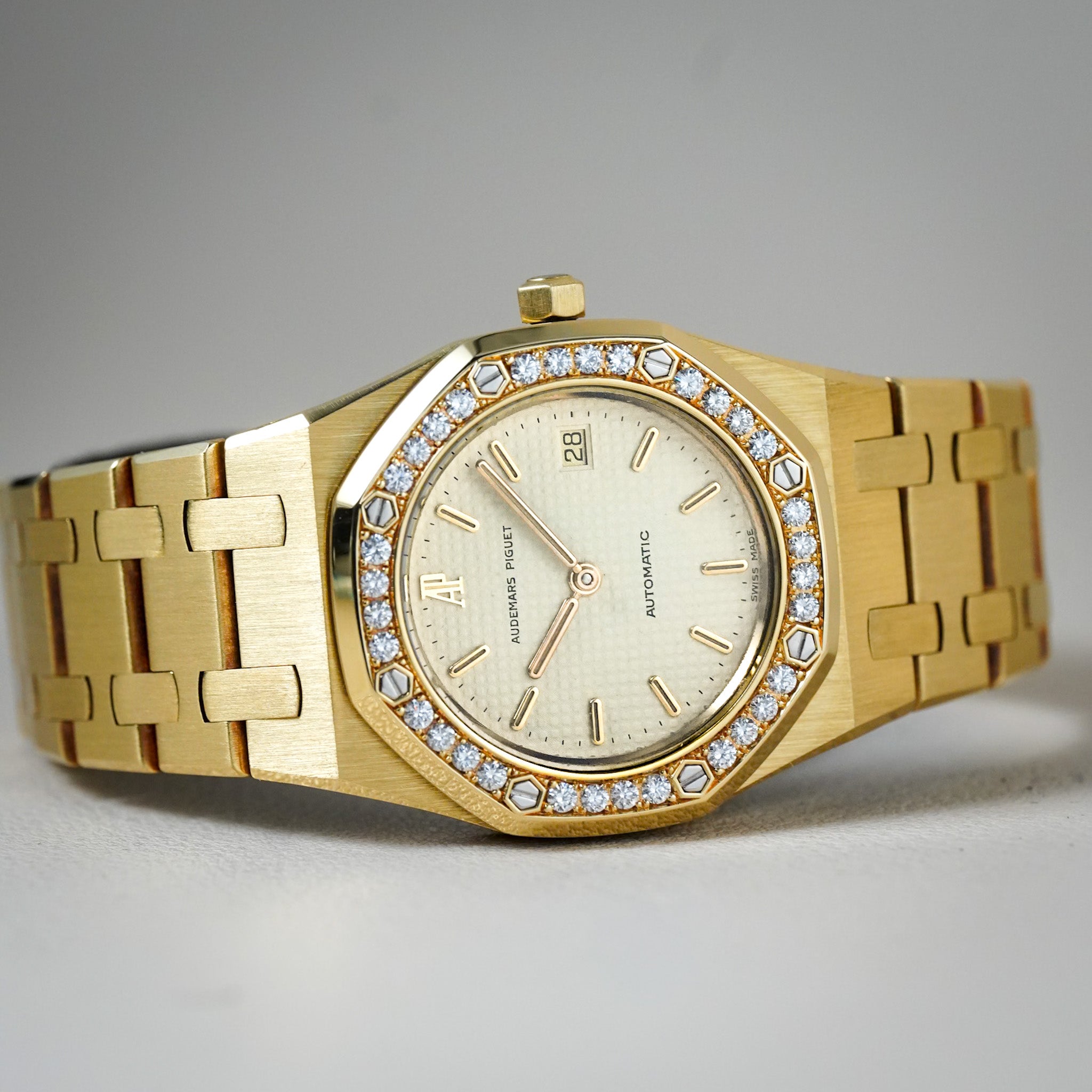 A gold-tone Audemars Piguet Royal Oak watch with a diamond-set bezel and a cream-colored dial, displayed on a neutral gray background.