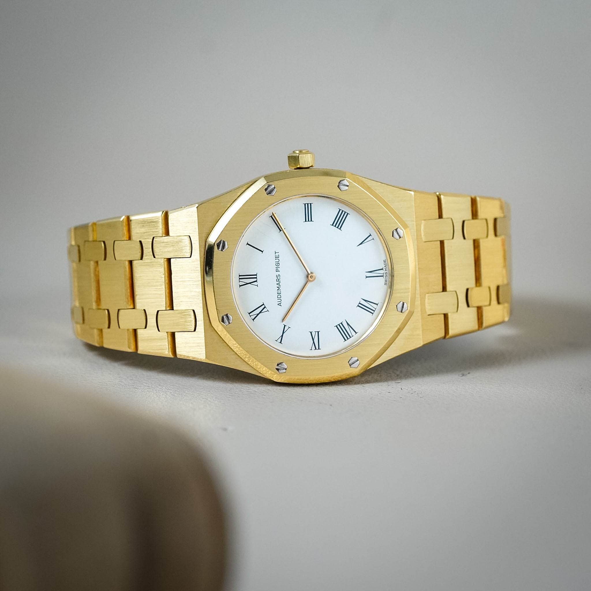 A gold-tone wristwatch with an octagonal bezel, white dial, and matching gold-tone link bracelet, photographed on a neutral light gray background.