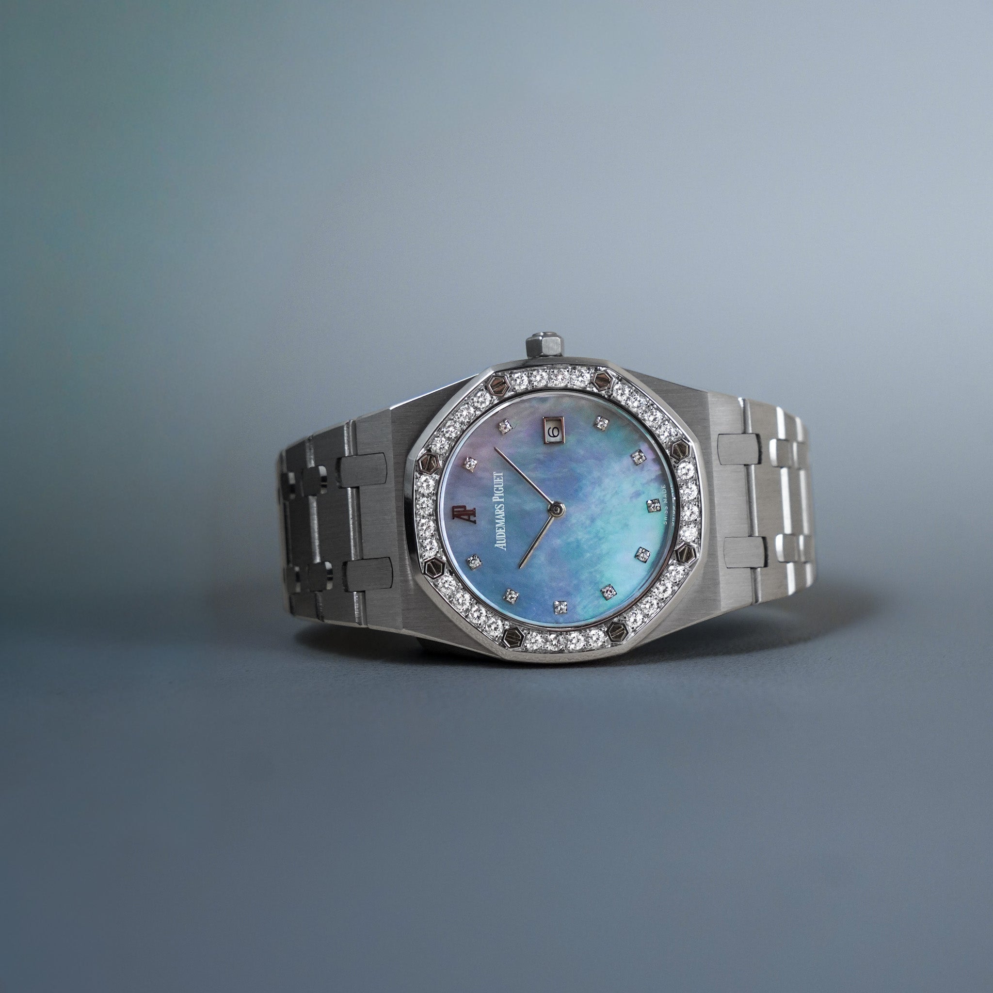 A luxury white gold wristwatch with a bezel-set diamond halo, a blue mother-of-pearl dial, and a matching metal link bracelet, displayed on a neutral gray background.