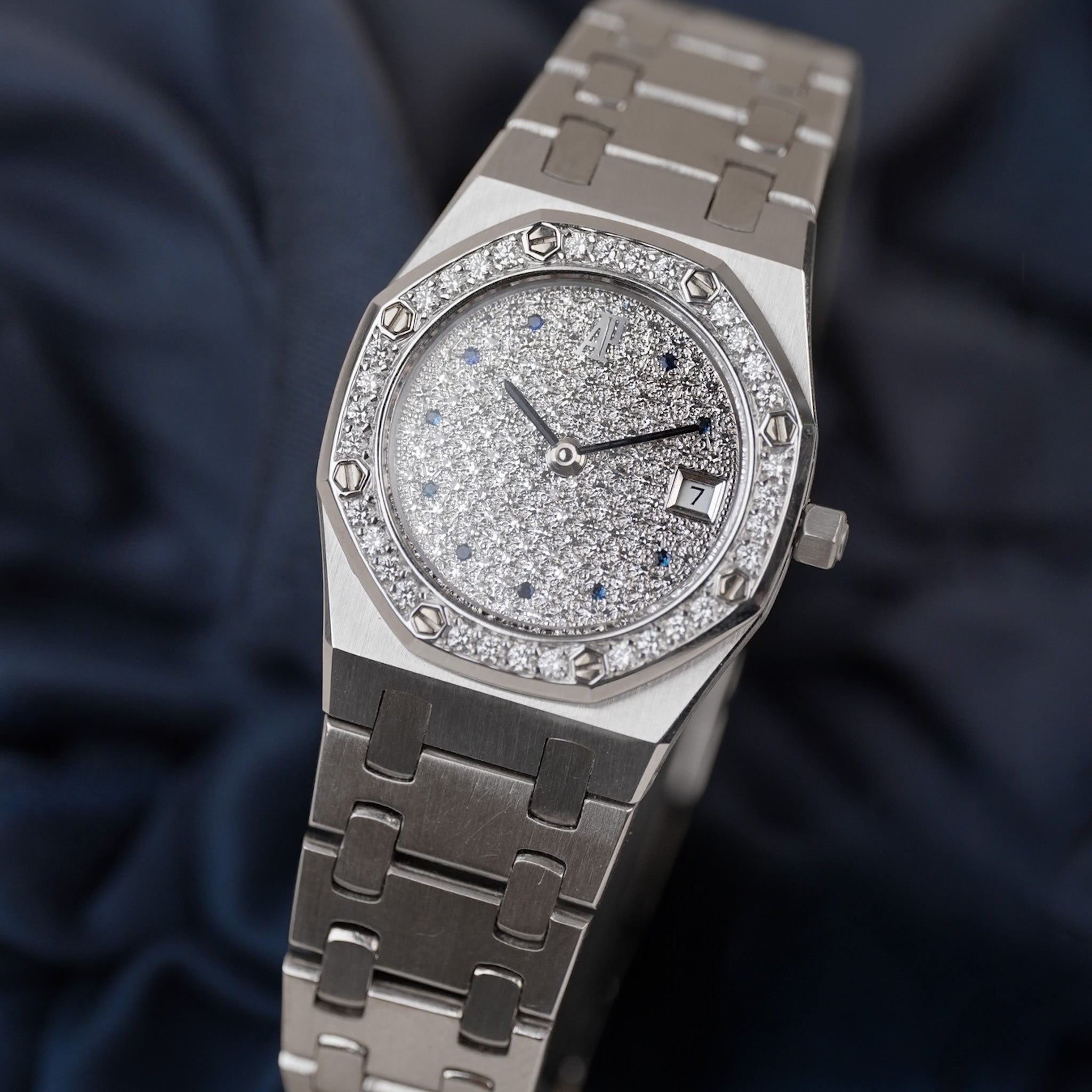 A luxury white gold stainless steel wristwatch with a diamond-encrusted bezel, a sparkling silver dial with blue accents, and a matching metal link bracelet, resting on a dark navy blue fabric background.