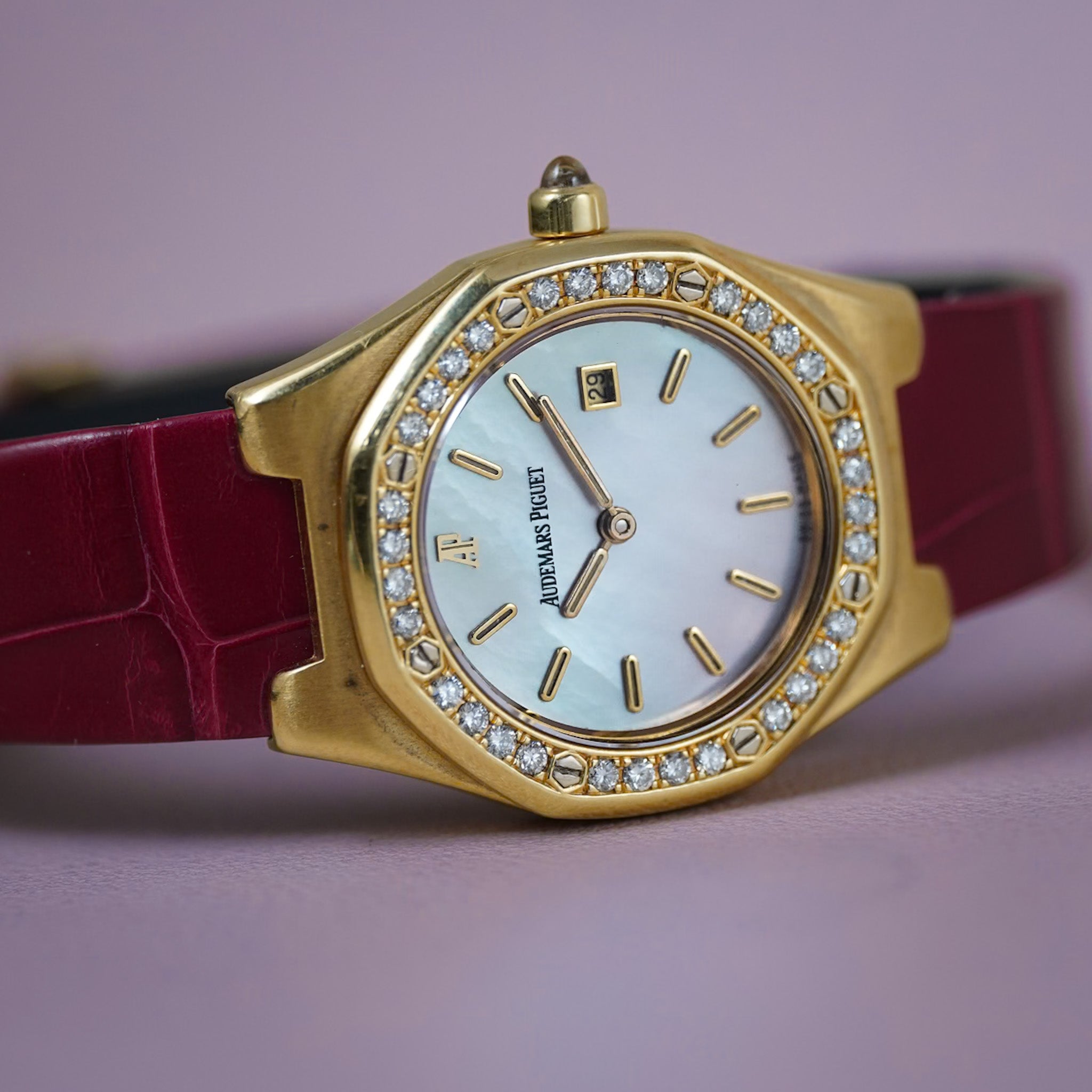 A yellow gold Audemars Piguet ladies watch with a diamond bezel, mother of pearl dial, and a red alligator leather strap, shot on a light purple background.