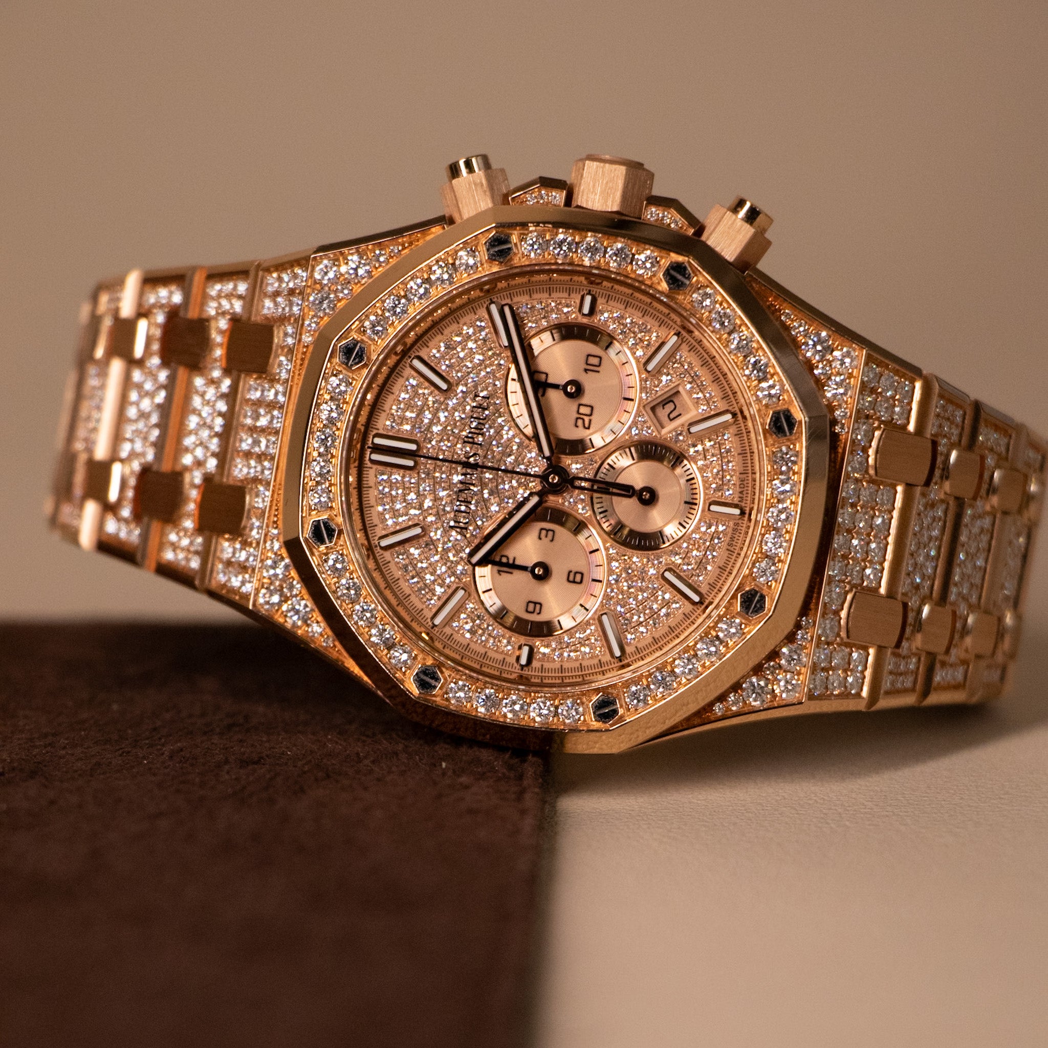 A luxury rose gold chronograph watch with a diamond-encrusted octagonal bezel and matching diamond-embellished bracelet, resting on a dark brown surface against a warm beige background.