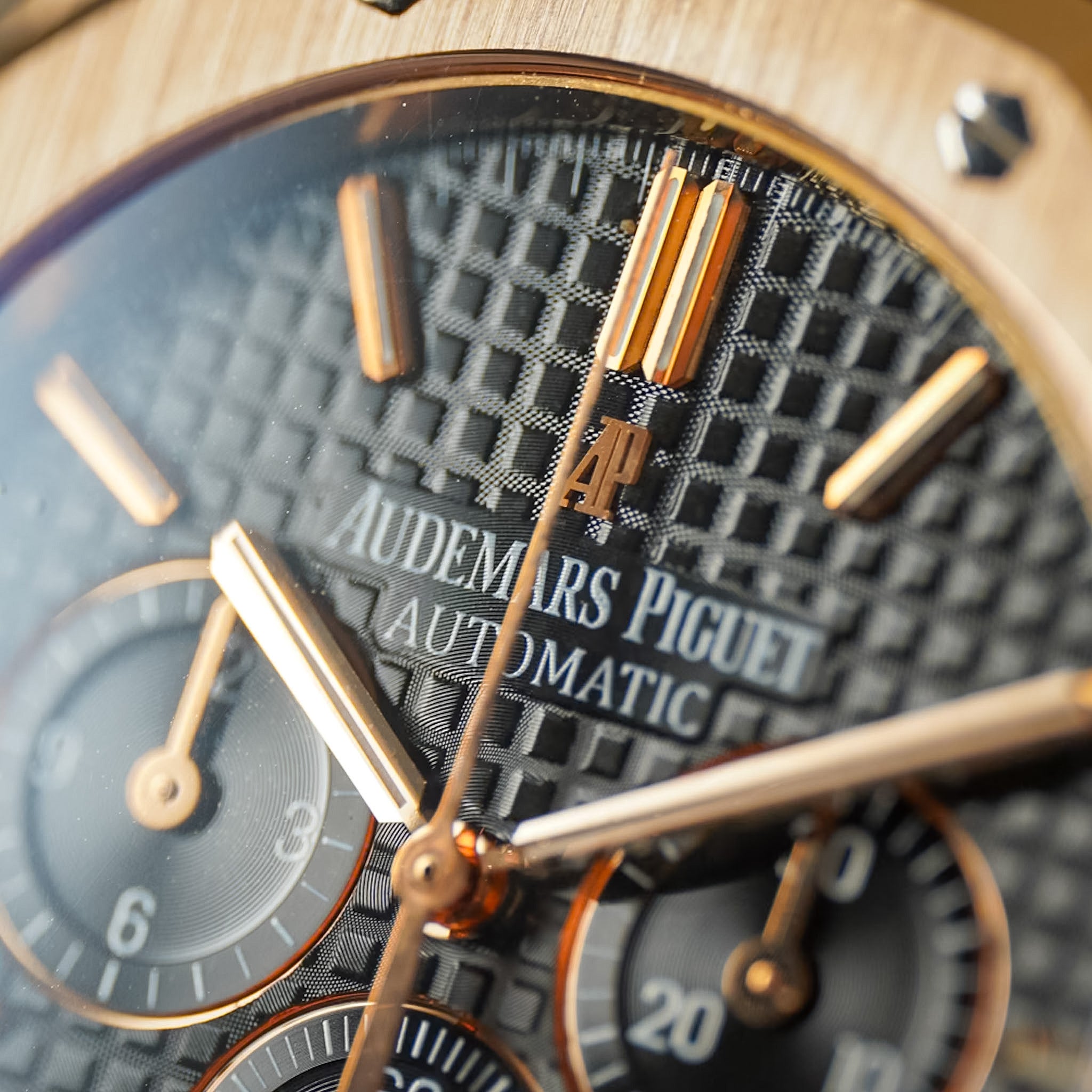Close-up of an Audemars Piguet Automatic watch dial with a textured black patterned face, rose gold-toned hands and markers, and a light wood-toned bezel.