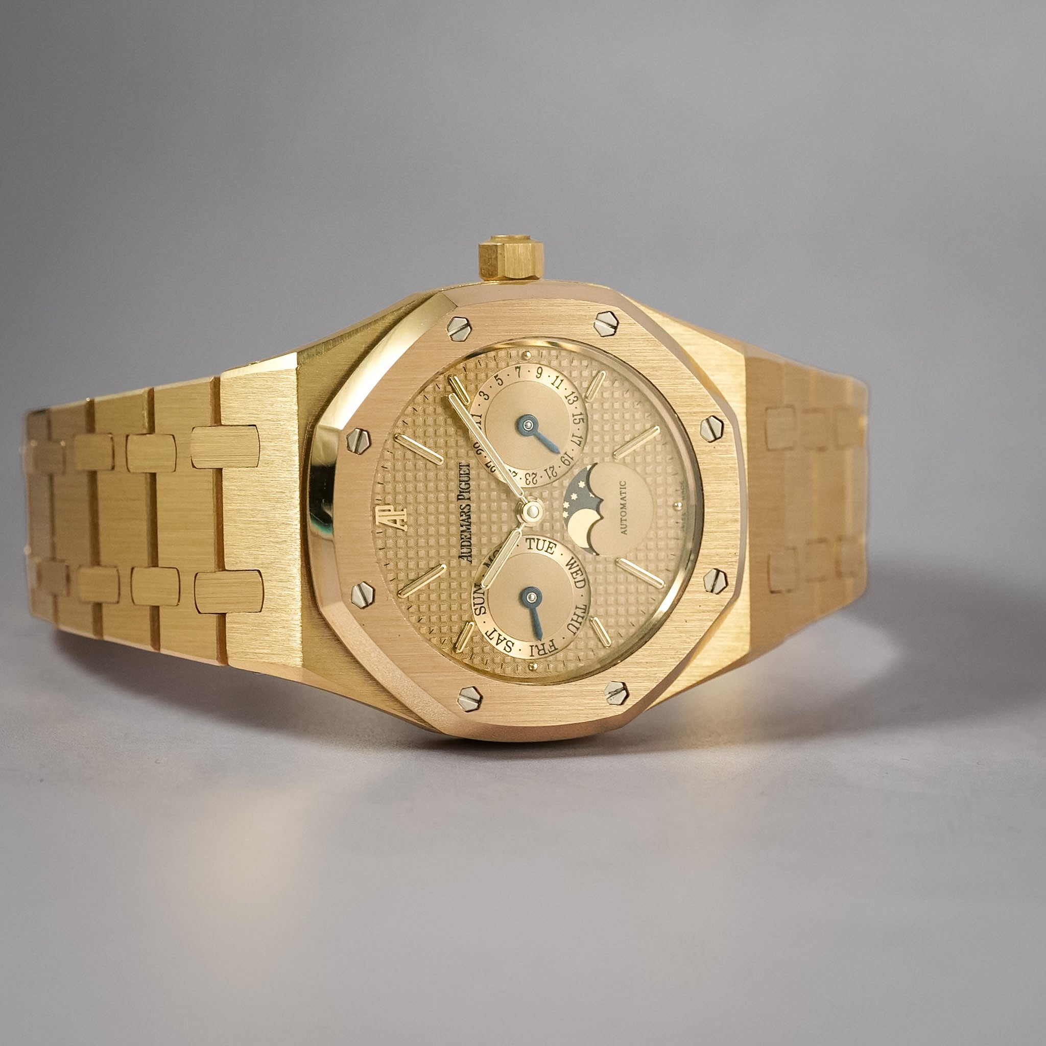 A yellow gold Audemars Piguet watch with a gold dial and three subdials laying sideways on a neutral grey background