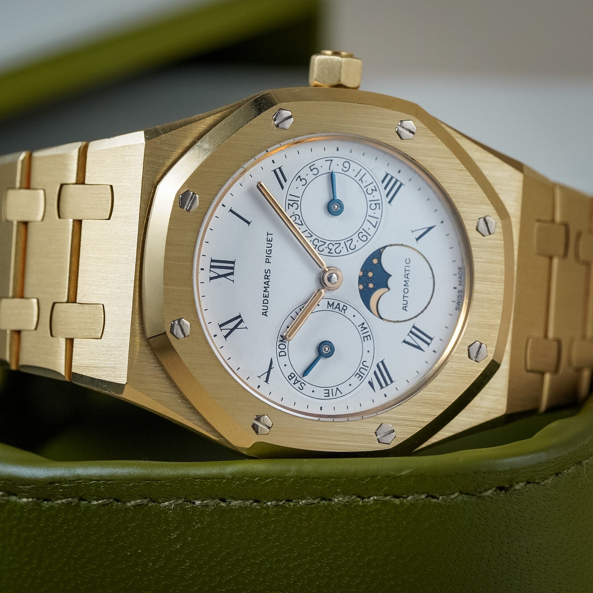 A close-up of a gold Audemars Piguet Royal Oak wristwatch with a white dial, Roman numeral hour markers, and a moon phase subdial, resting on an olive green leather box.