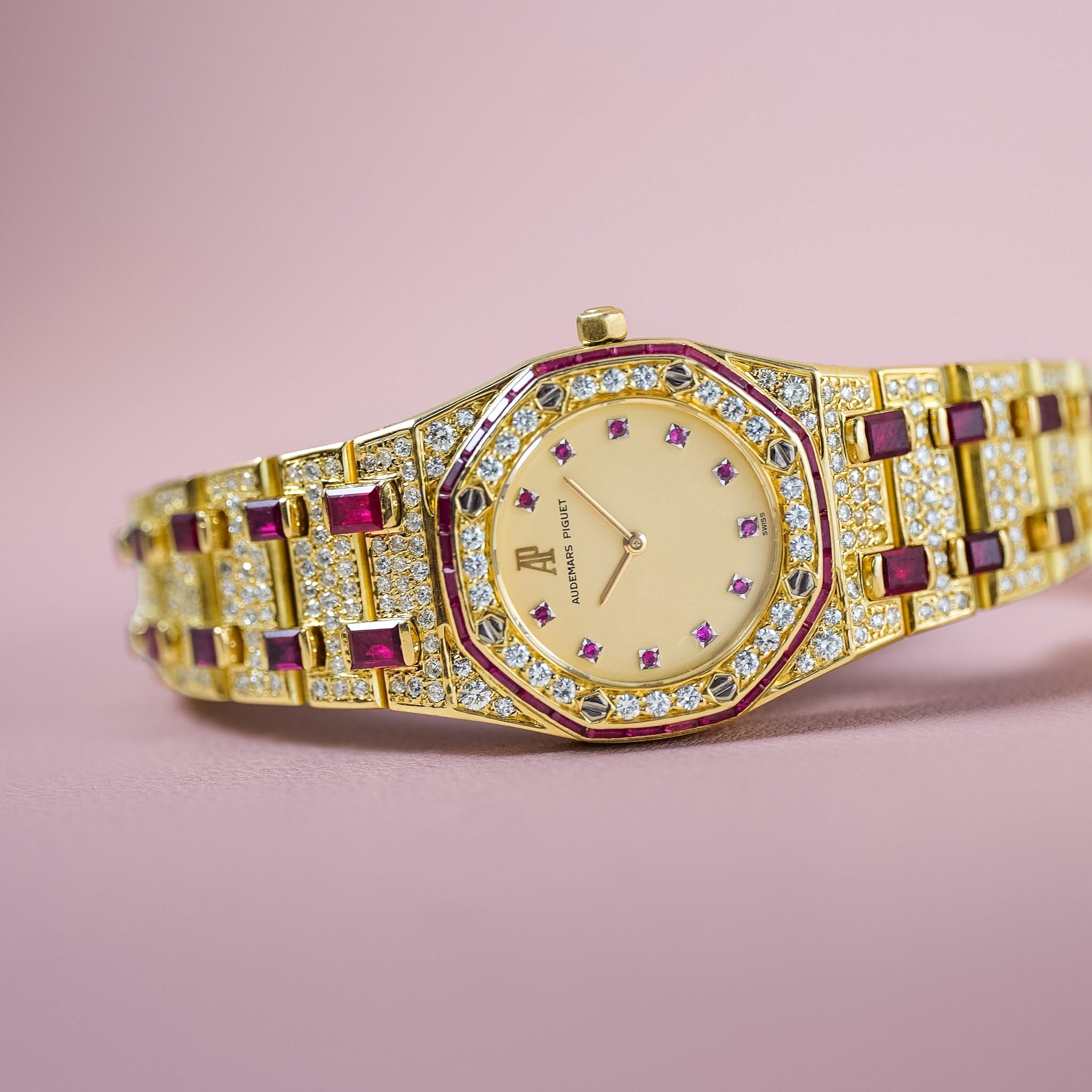 A gold-tone wristwatch with a diamond-encrusted bracelet, featuring deep pinkish-red round gemstones and a gold dial with diamond hour markers, set against a solid pink background.