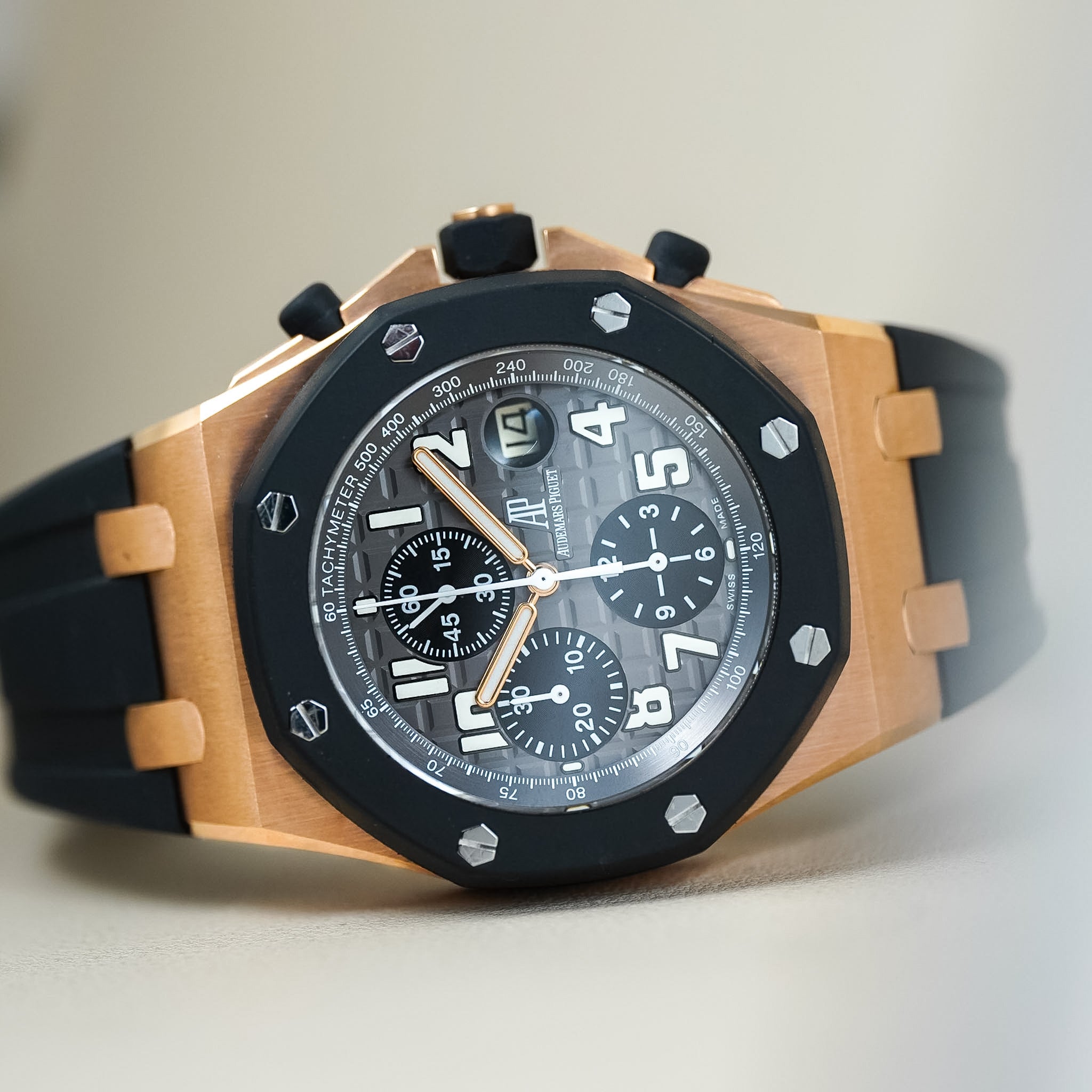 An Audemars Piguet Royal Oak Offshore Chronograph with a rose gold case, black ceramic bezel, textured dial with chronograph subdials, and a black rubber strap, laying on its side on a neautral cream background