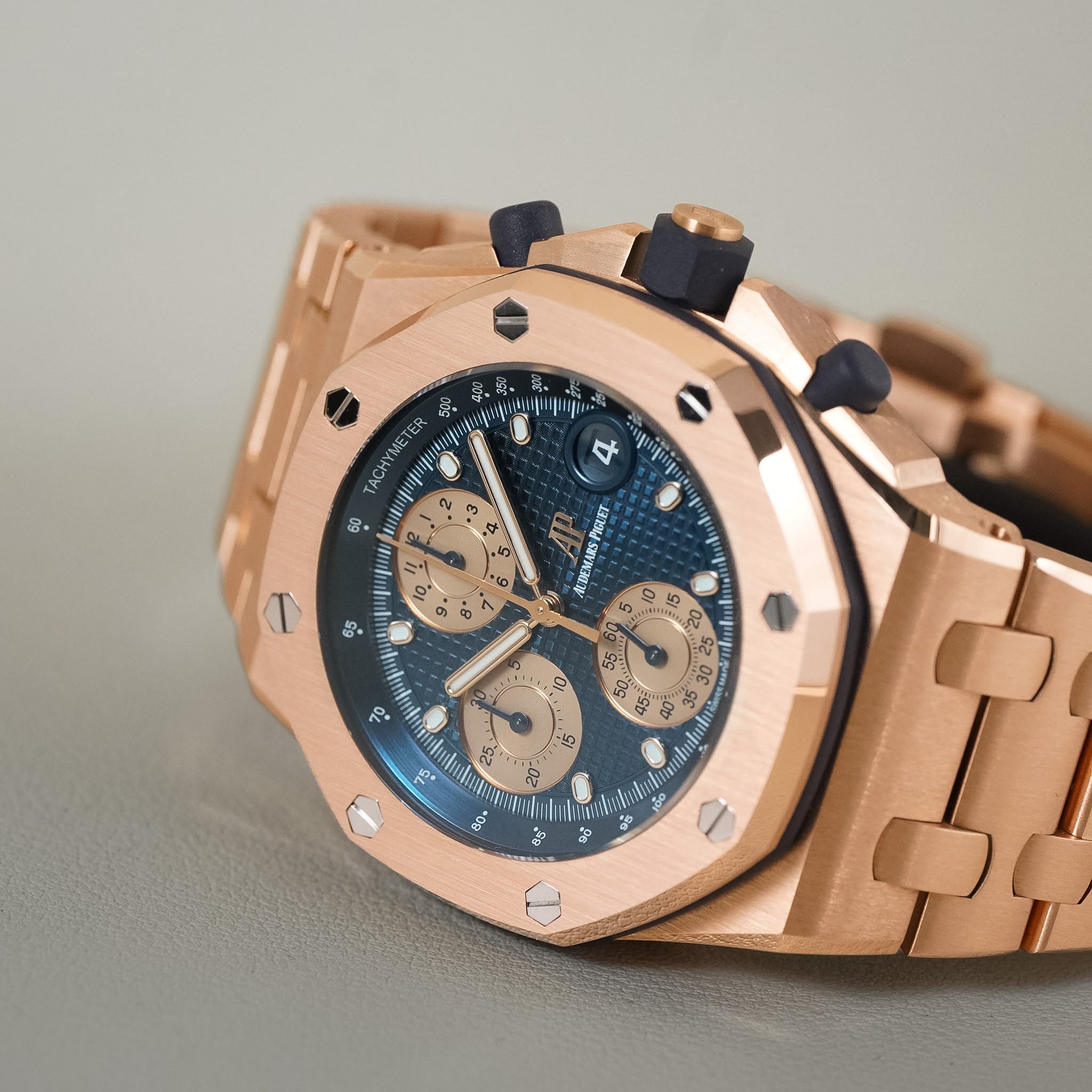 A close-up shot of an Audemars Piguet Royal Oak watch featuring a rose gold case, a blue textured dial with beige subdials, and a matching rose gold bracelet, set against a neutral gray background.