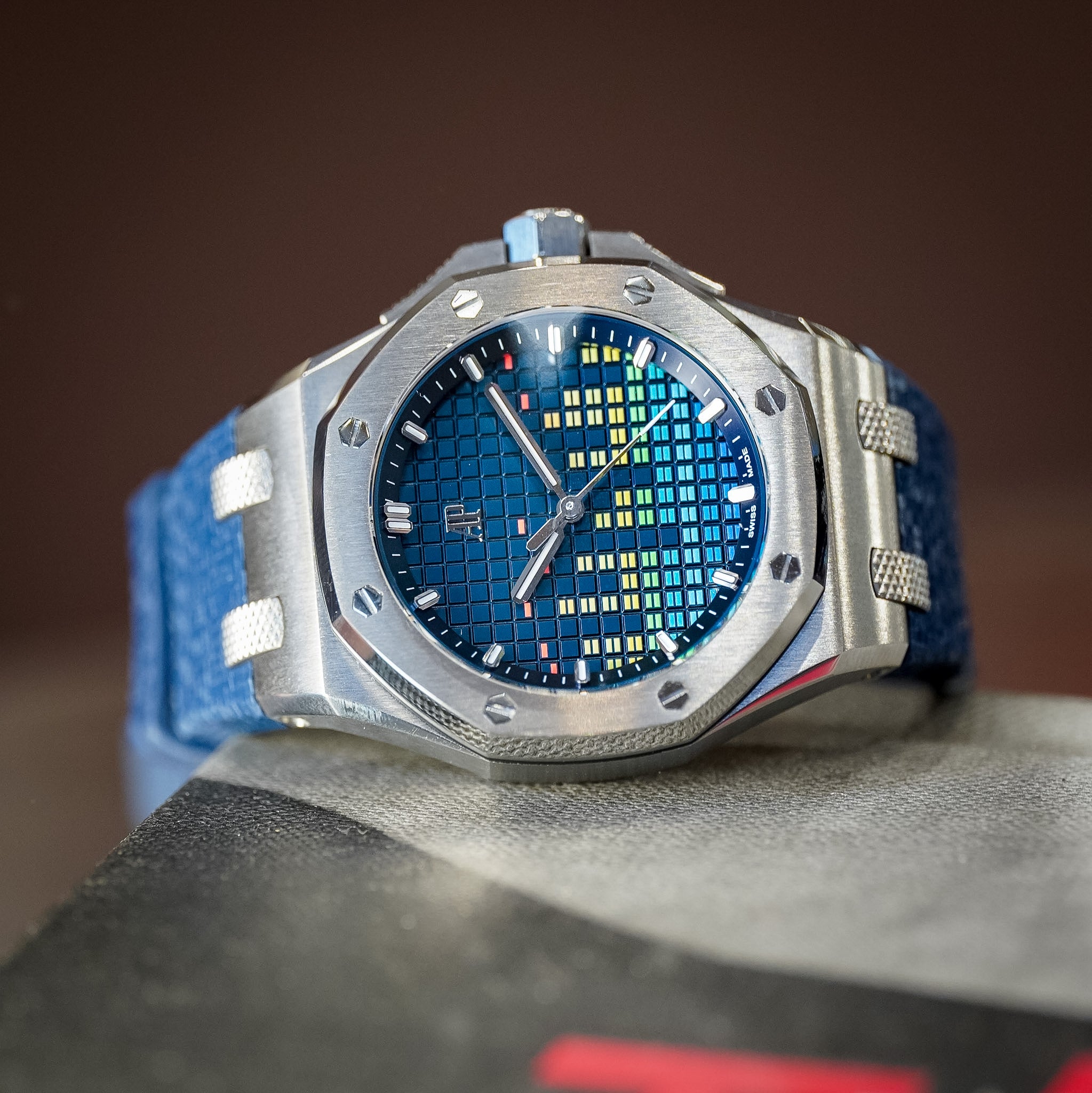 A luxury wristwatch with a silver-toned octagonal case, a textured blue dial with a multi-colored equalizer motif, and a blue textured strap, resting on a gray surface against a dark brown background.