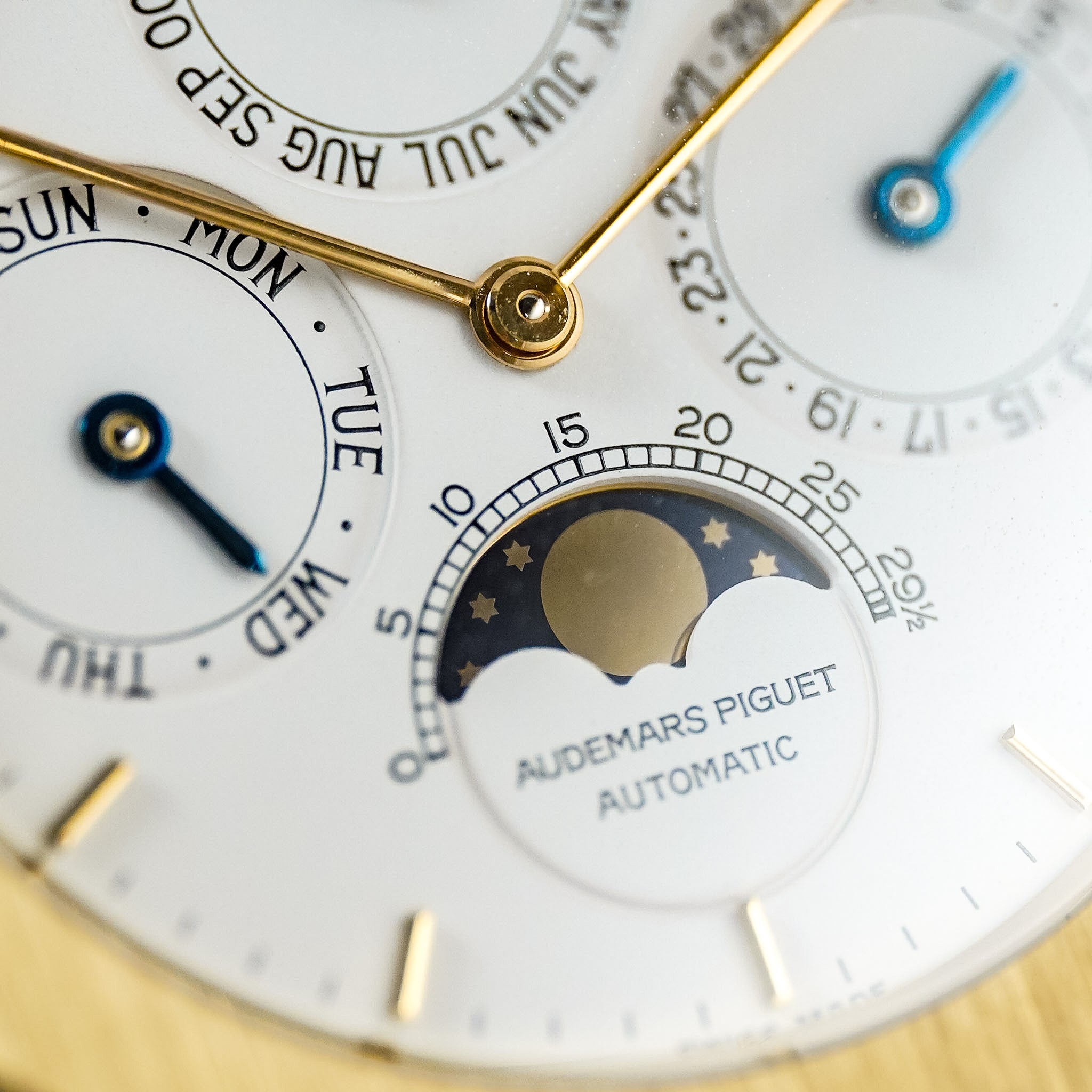 Close-up of an Audemars Piguet Automatic watch dial, showing white subdials, gold hands and markers, a moon phase window, and blue accent details.