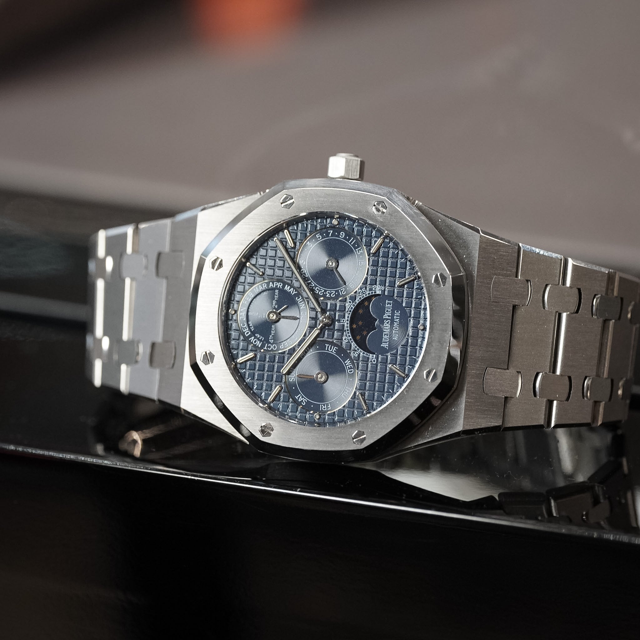 A luxury stainless steel wristwatch with an octagonal bezel, blue textured dial, and matching blue subdials, resting on a glossy black surface.