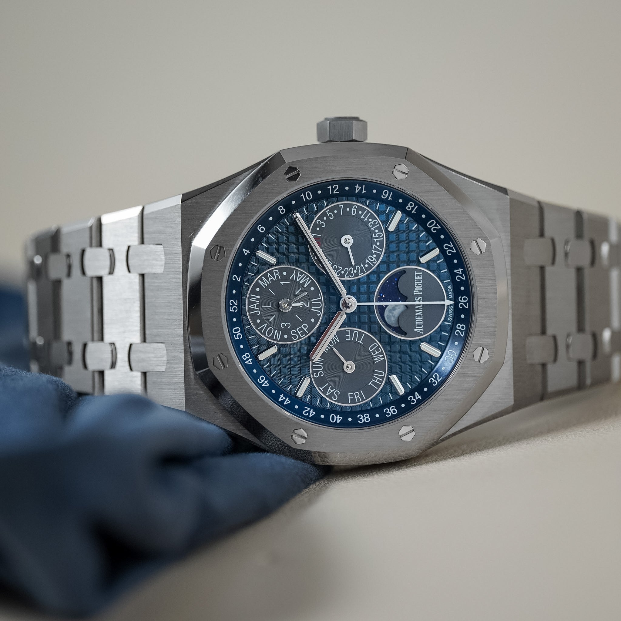 A luxury wristwatch with a brushed silver-tone metal case and integrated link bracelet, featuring a textured blue dial with multiple subdials and a silver bezel, resting on a light beige surface with a blue cloth in the foreground.