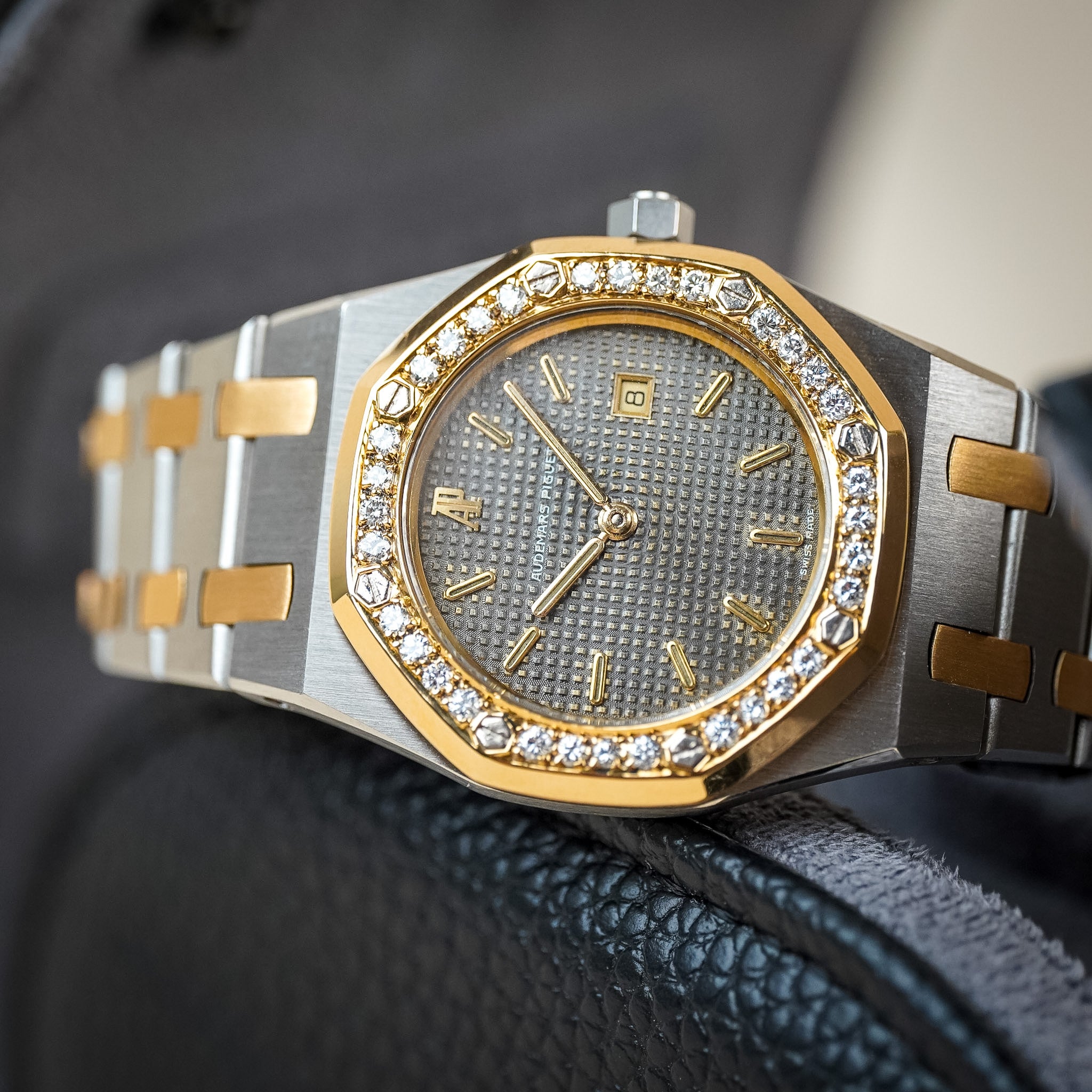 A luxury two-tone stainless steel and yellow gold wristwatch with a diamond bezel and textured gray dial, resting on a black leather surface.