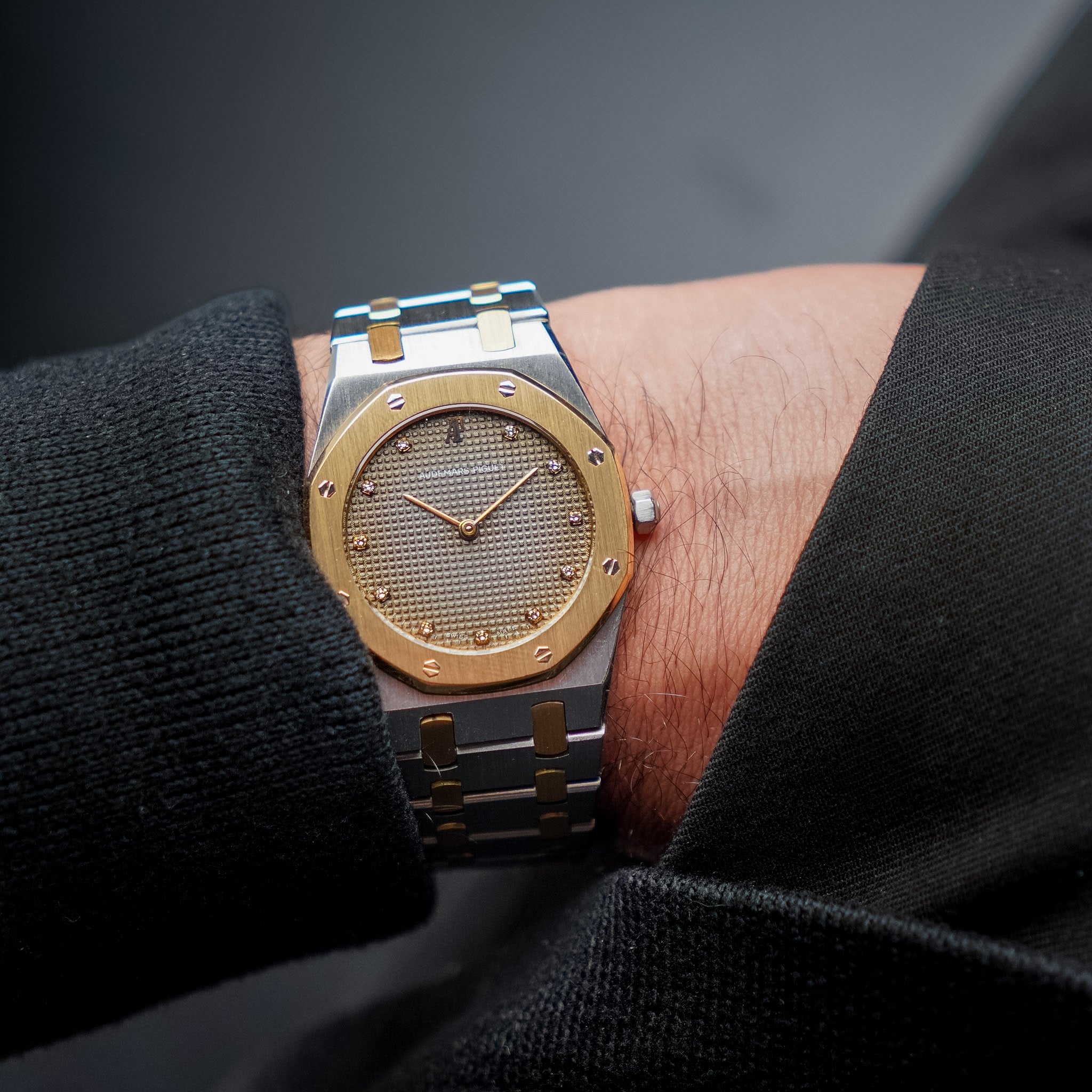 A man's wrist in black clothing wearing a stainless steel and yellow gold Audemars Piguet Royal Oak with a faded grey dial with diamond hour markers.