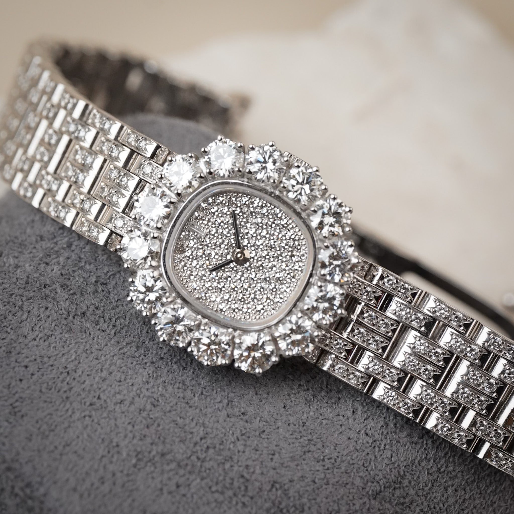 A diamond-encrusted silver-tone watch with a square bezel and matching pavé dial, displayed on a dark gray velvet surface with a soft beige background.